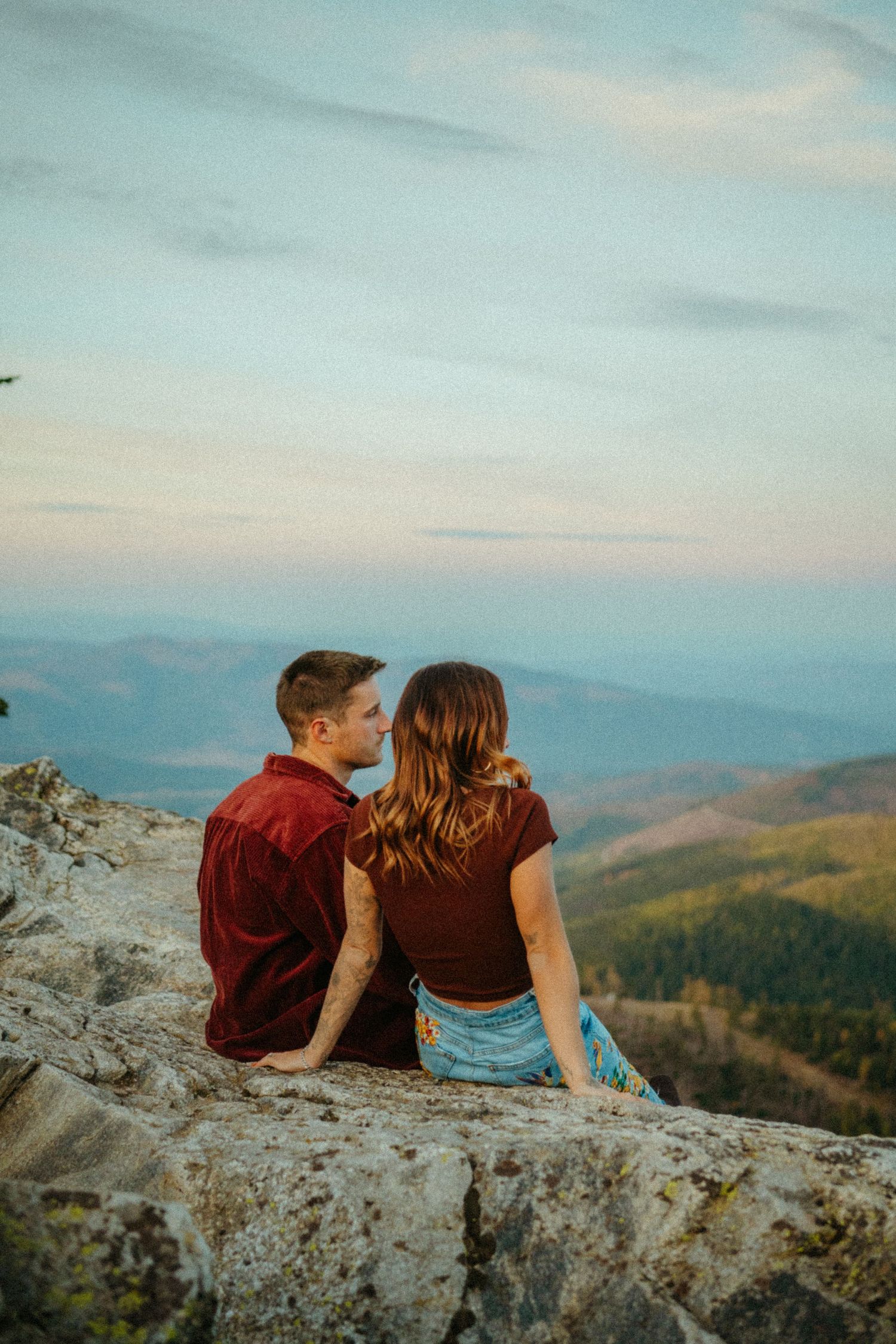 Couple in matching burgundy shirts sits together on mountain cliff overlooking scenic valley at sunset.