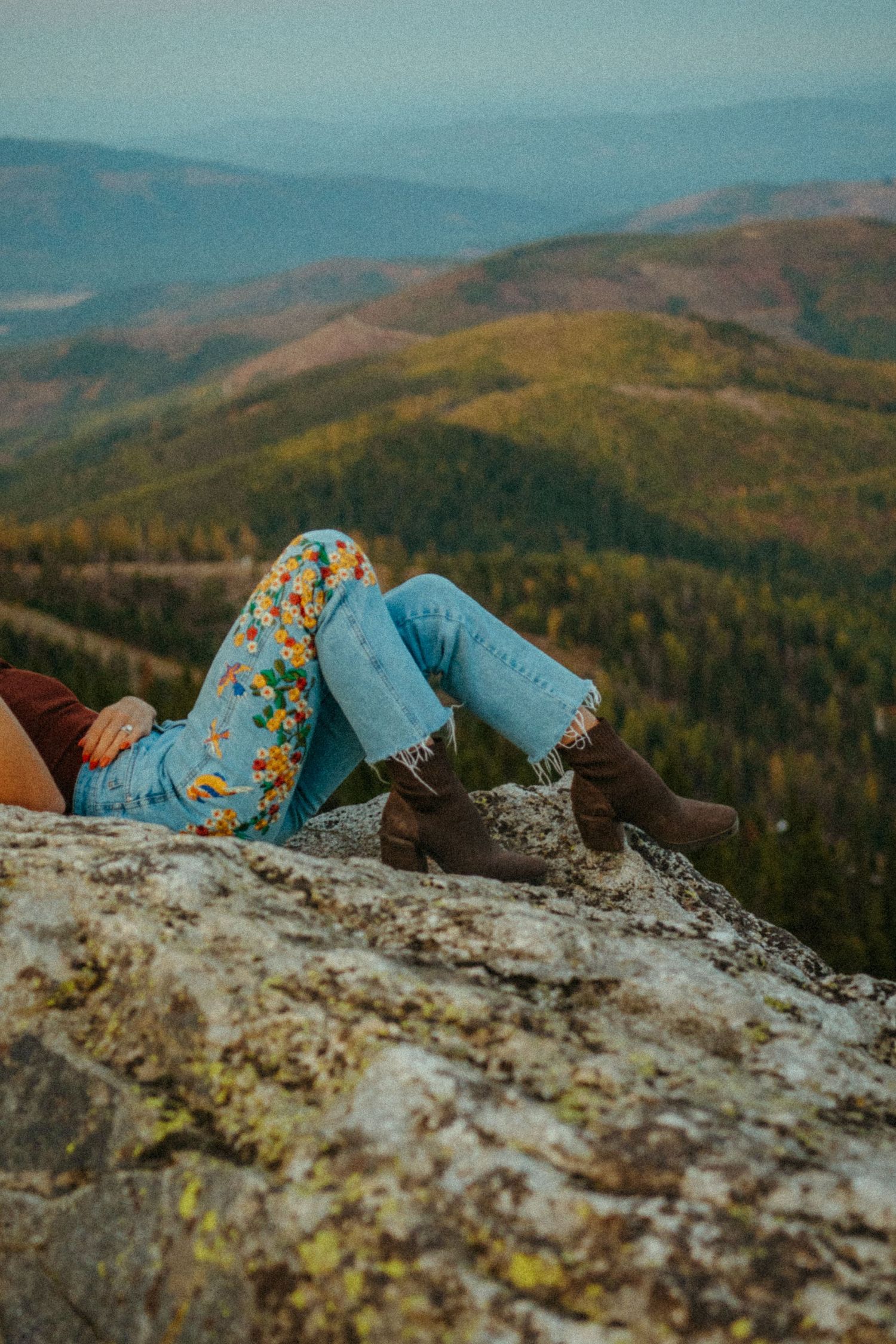 Someone in boots and jeans with floral pattern clothing reclines on a mountaintop overlooking a vast autumn forest landscape.