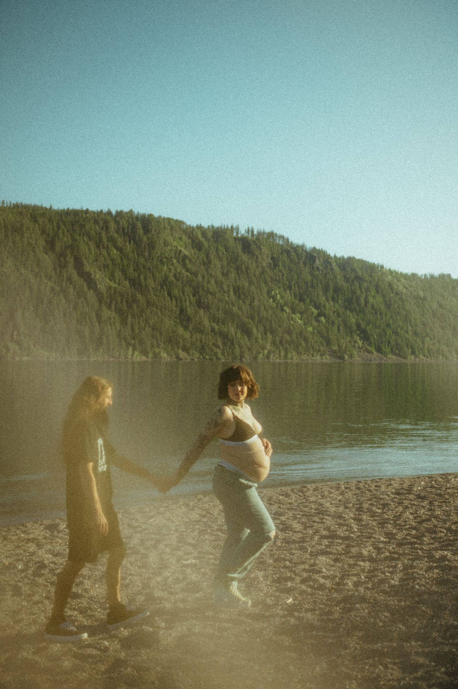 Two people enjoying a sunny afternoon on a lakeside beach with forested hills in the background.