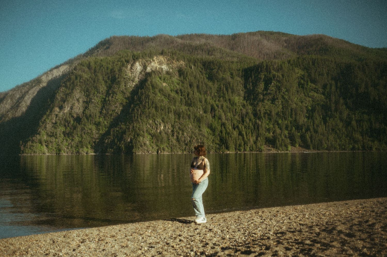 Tranquil lakeside scene with a figure on sandy beach surrounded by forested mountains in natural lighting.