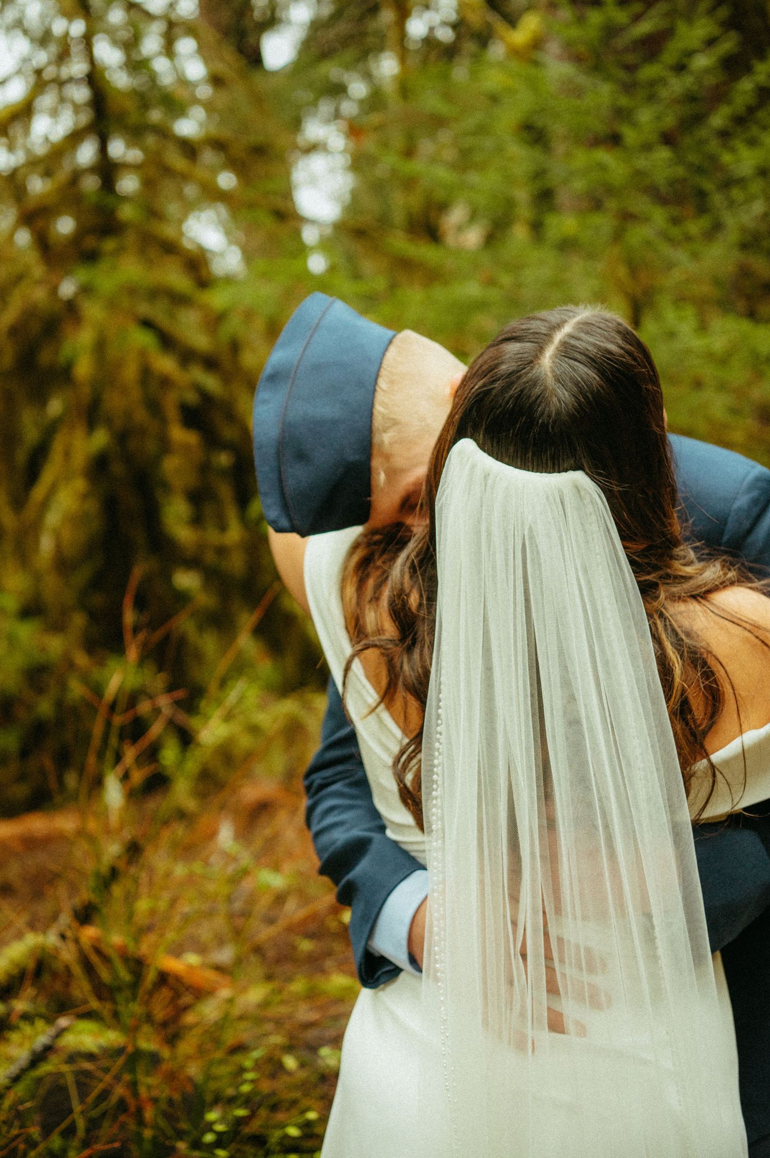 A romantic wedding moment captured in a forest setting with a flowing white veil against greenery in the background.