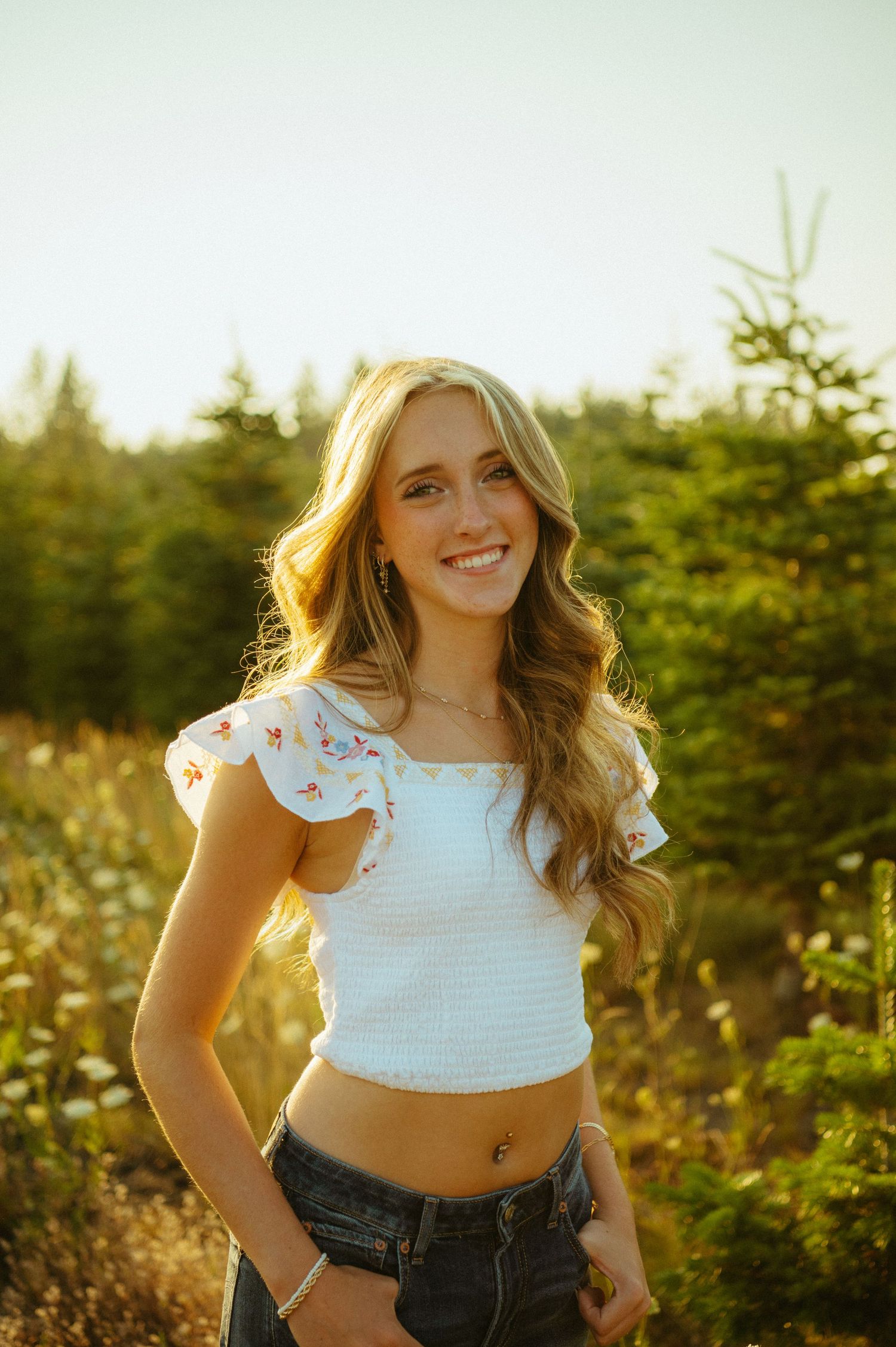 A person in a white crop top and denim shorts poses outdoors during golden hour with sunlight filtering through trees.