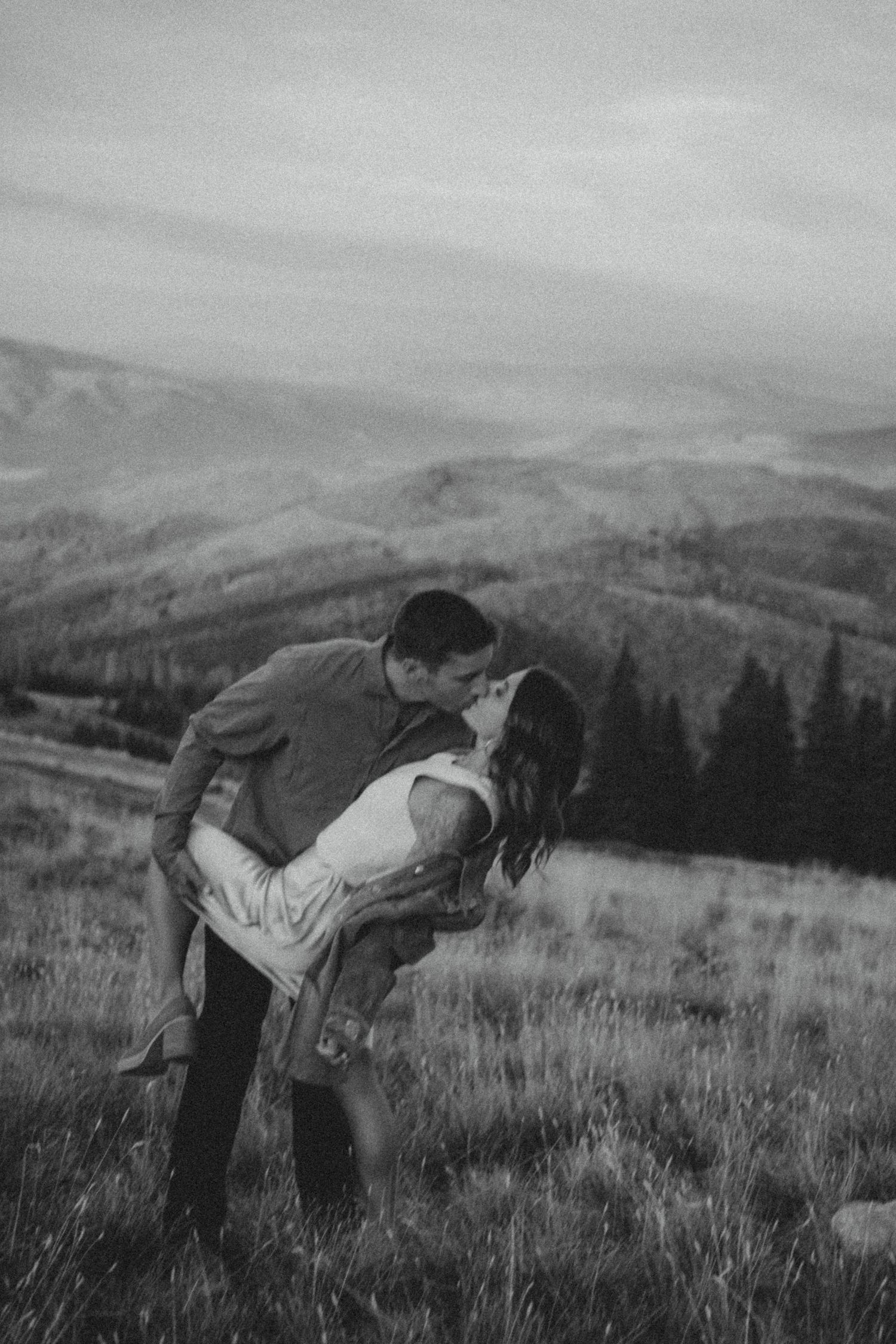 A romantic black and white scene shows a couple sharing a passionate embrace on a hilltop overlooking a mountain landscape.