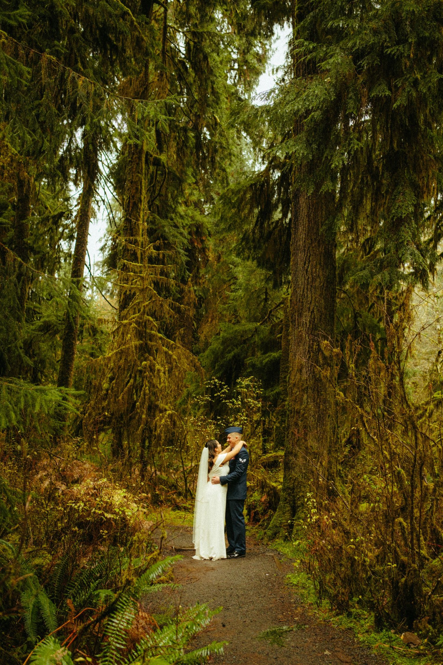 A couple embraces on a misty forest path surrounded by towering evergreen trees draped in moss in the Pacific Northwest.
