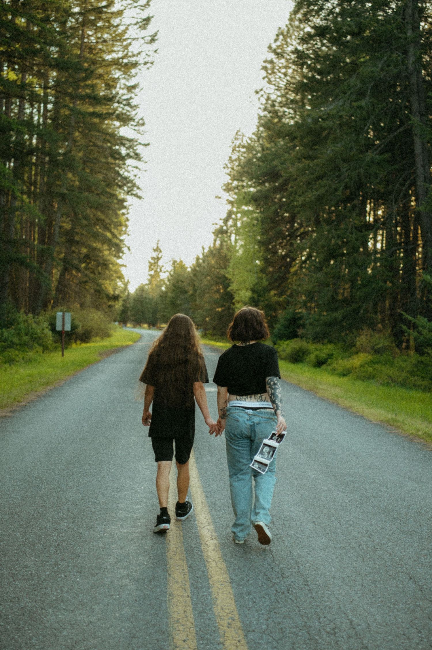 Two friends walk together holding hands on a winding forest road lined with tall evergreen trees at dusk.