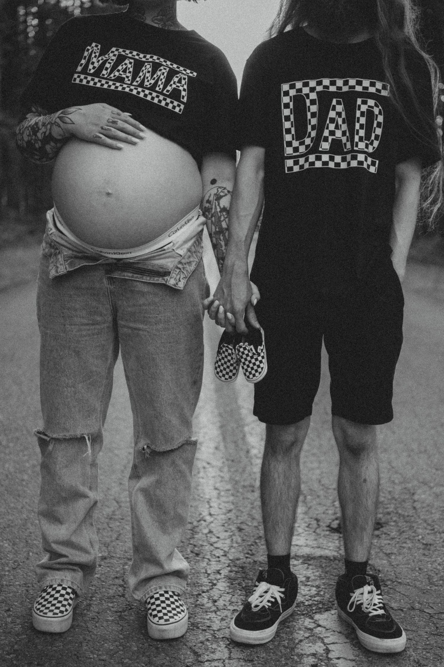 Two people wearing matching checkered Vans shoes stand together, one showing a pregnant belly in black and white photo.
