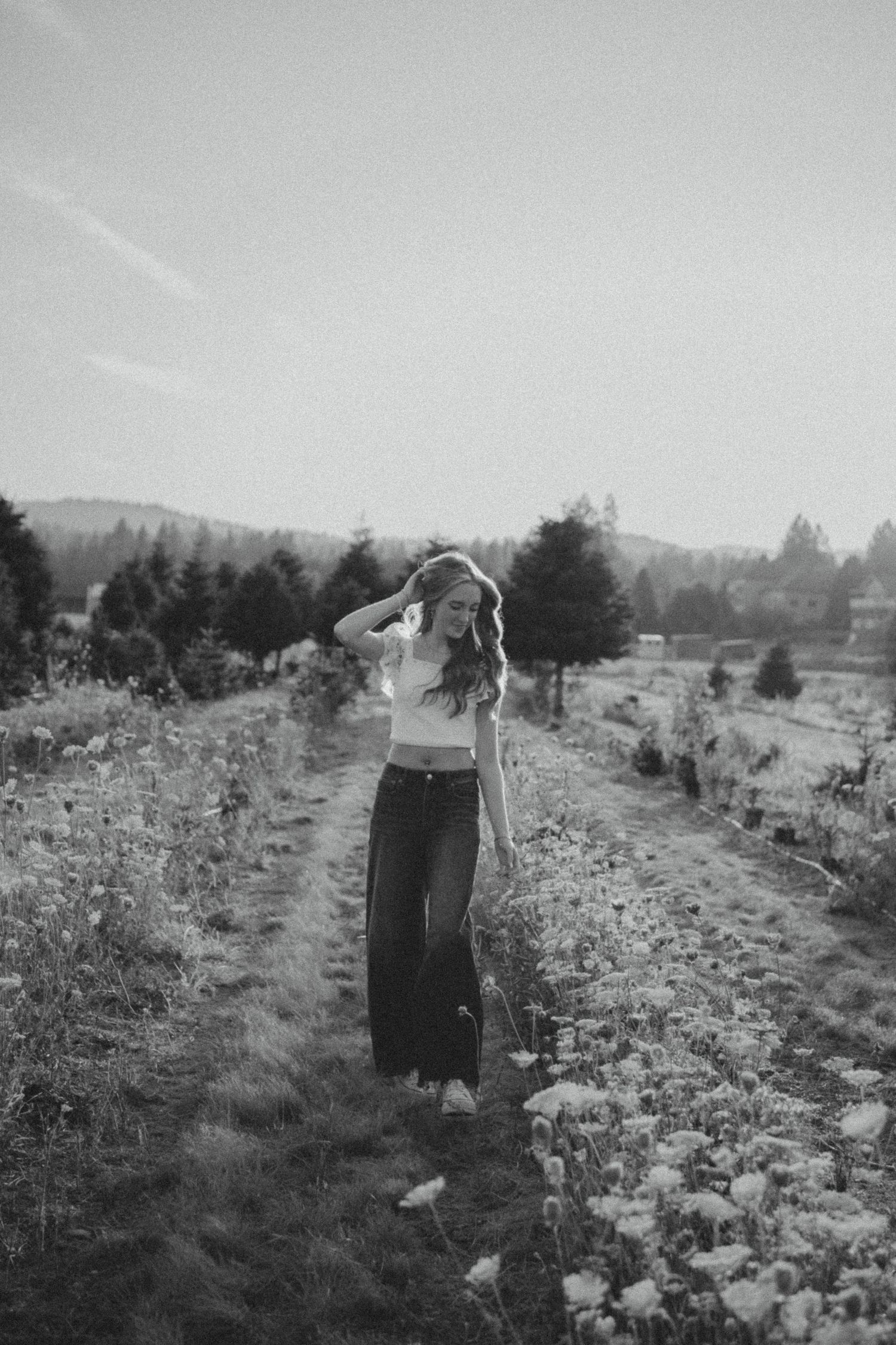 Black and white artistic landscape photo showing a person walking on a rural path surrounded by wildflowers and trees.