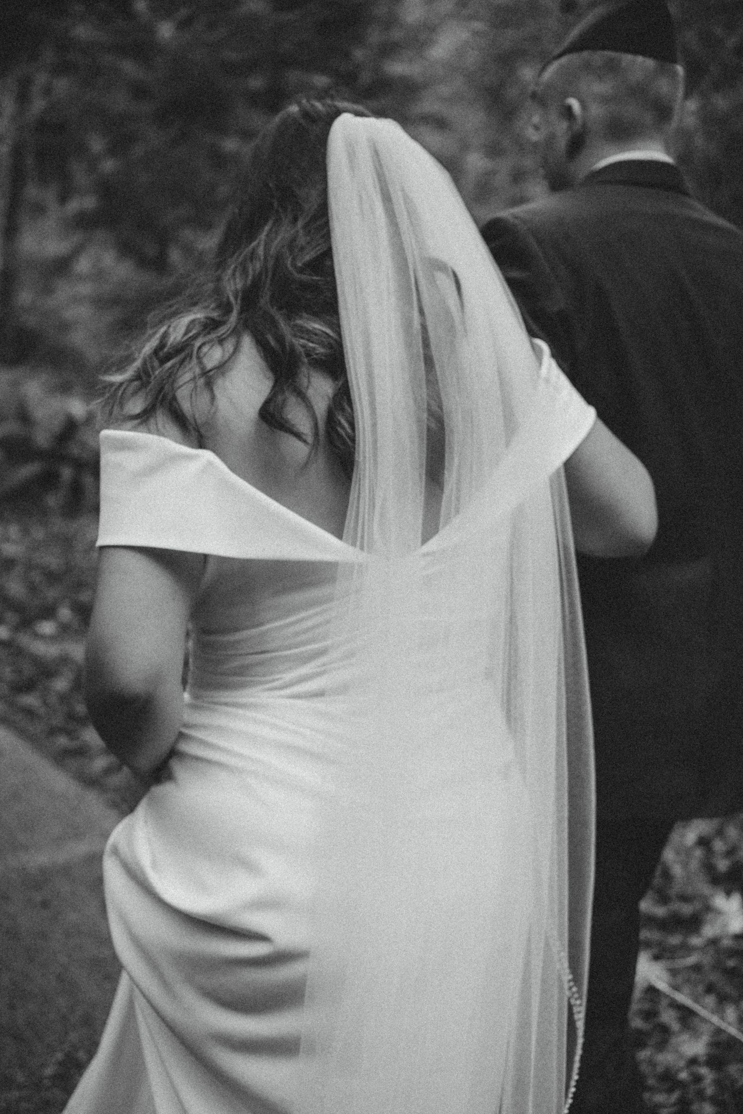 Black and white candid wedding photo showing back view of bride in elegant white dress with flowing veil.