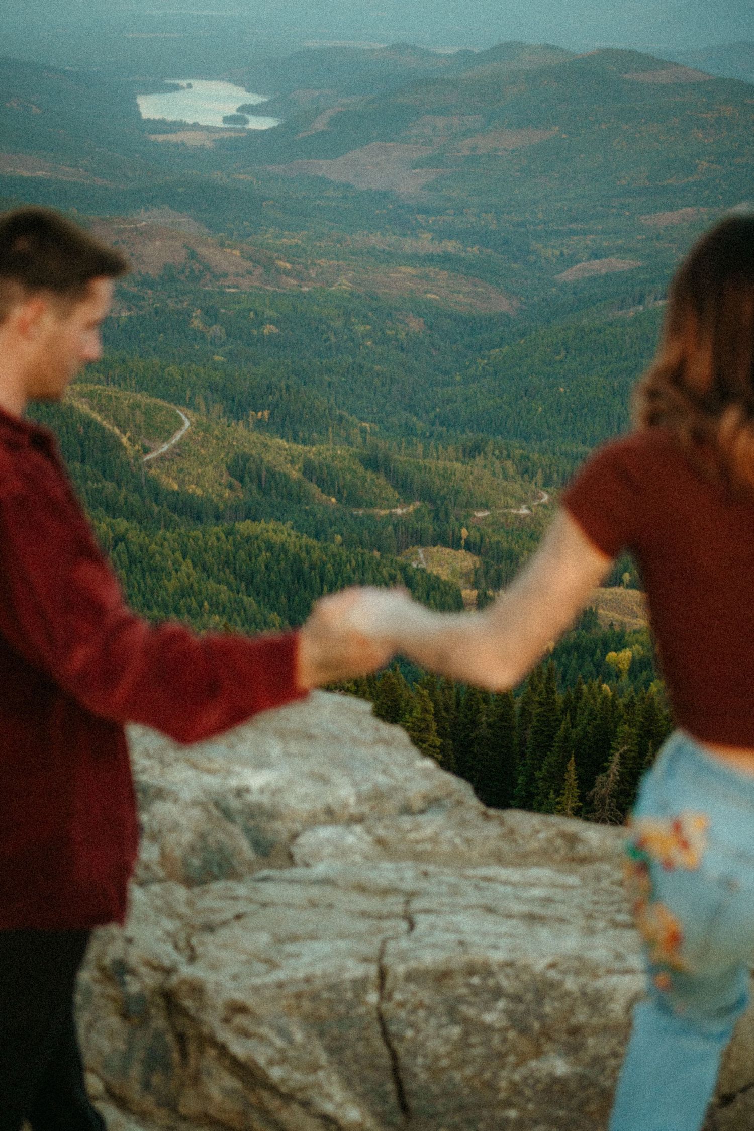 Couple holds hands on mountain overlook with scenic lake and forested hills stretching into misty distance.