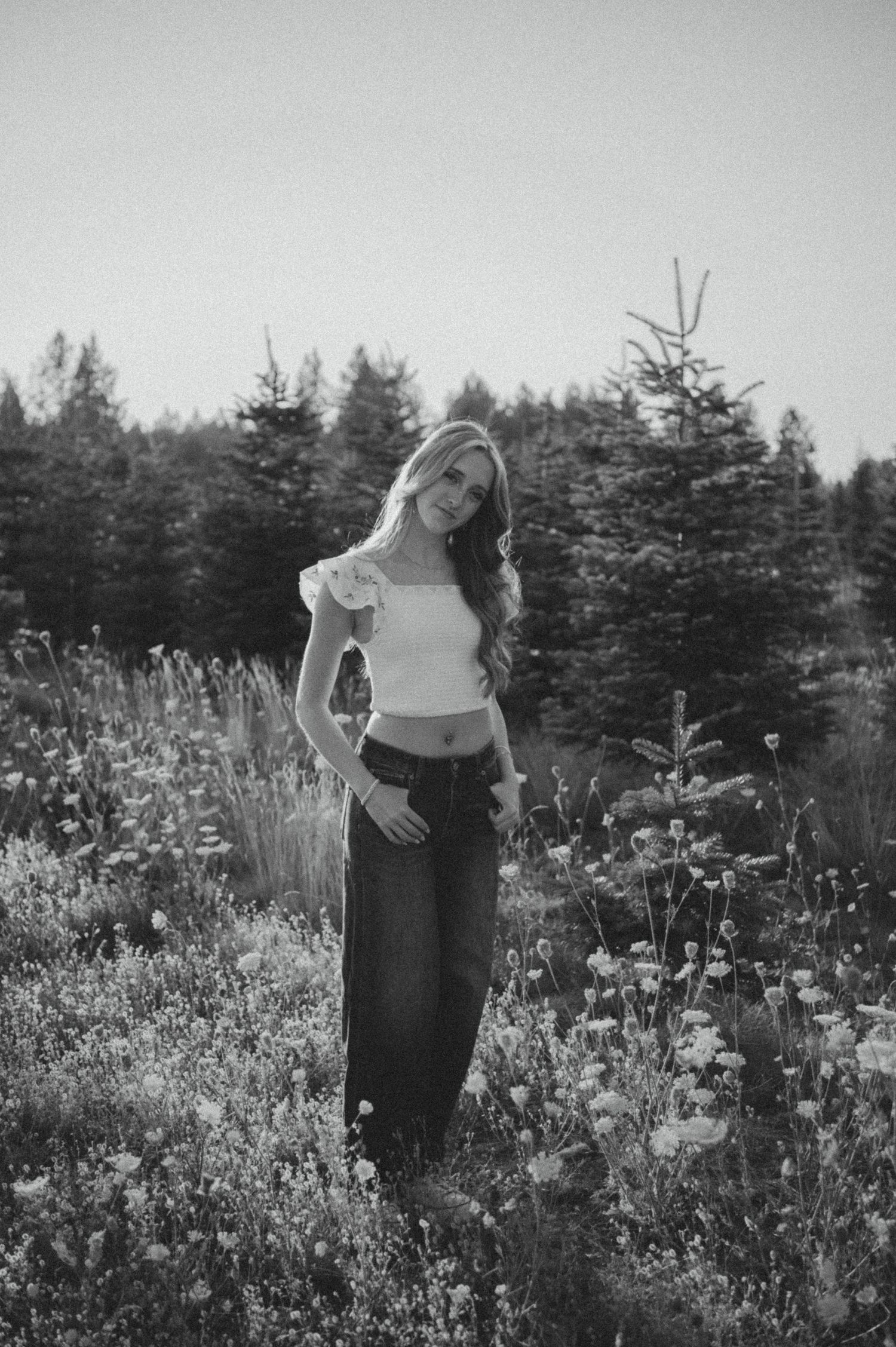 A black and white photo shows a figure standing in a wildflower field surrounded by evergreen trees.