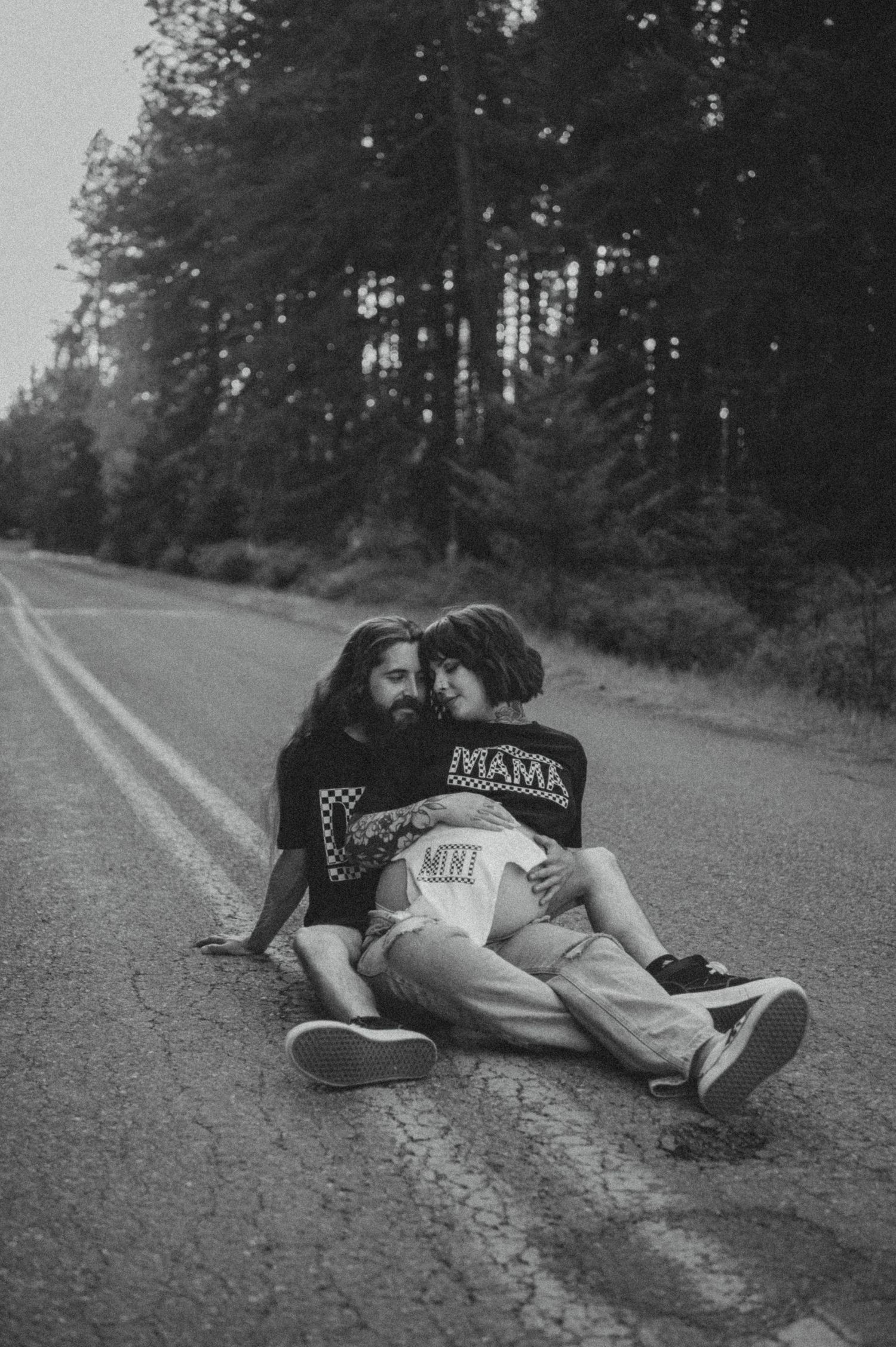 A couple sits intimately on an empty rural road with pine trees in the background, captured in black and white.