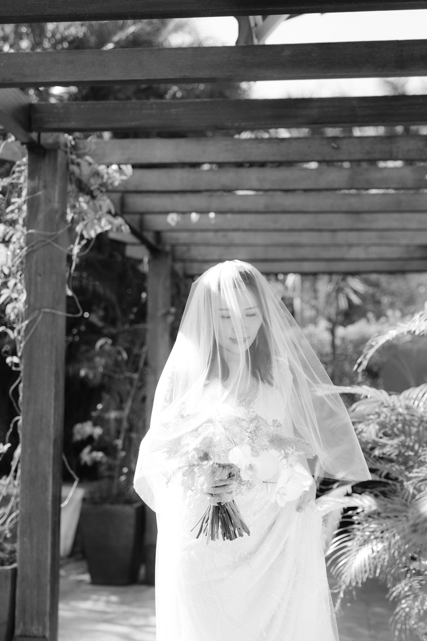 Black and white photo of a bride holding a bouquet in an outdoor garden setting with wooden pergola overhead.