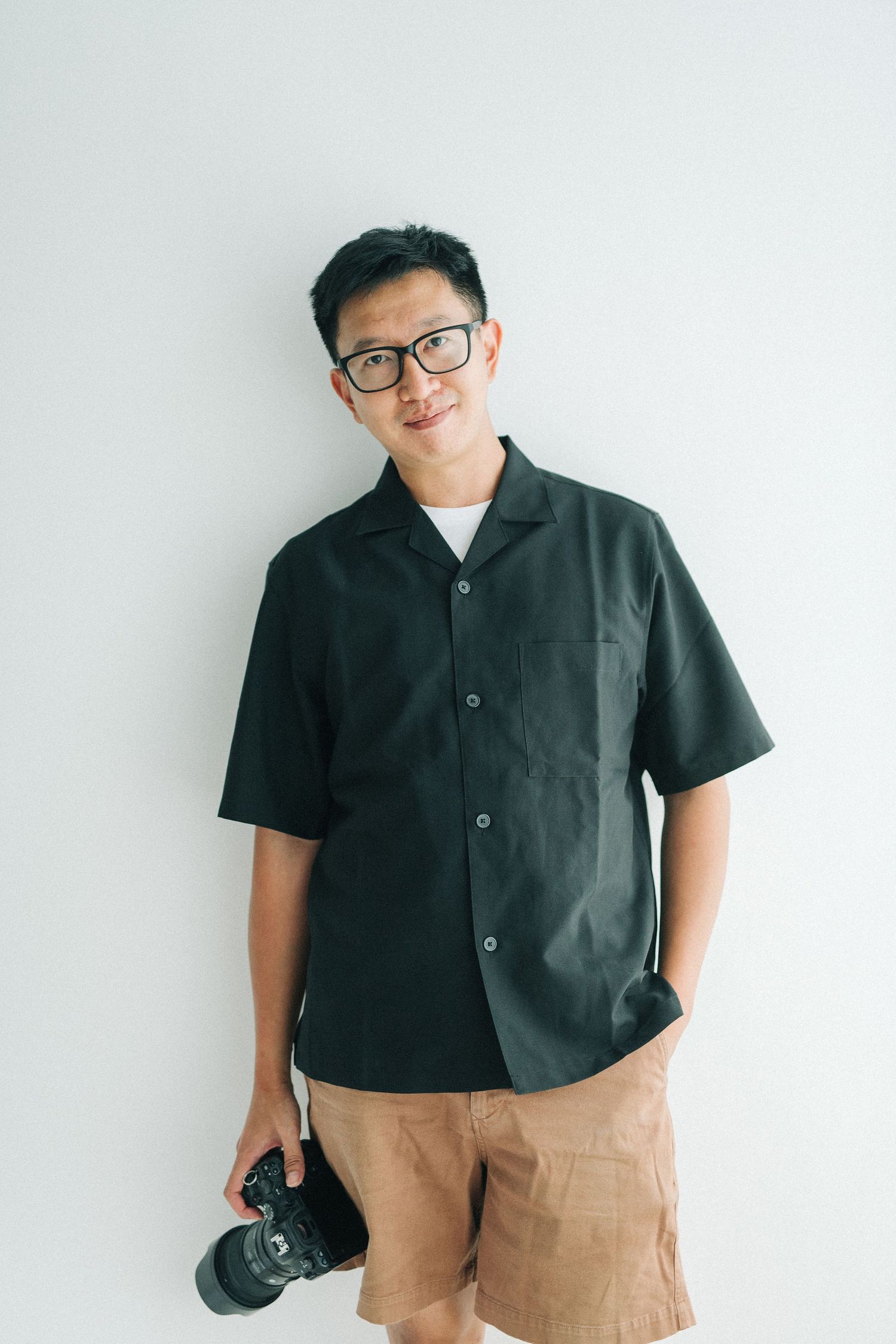 Young Asian man in black shirt and glasses holding a camera, smiling against a white background.