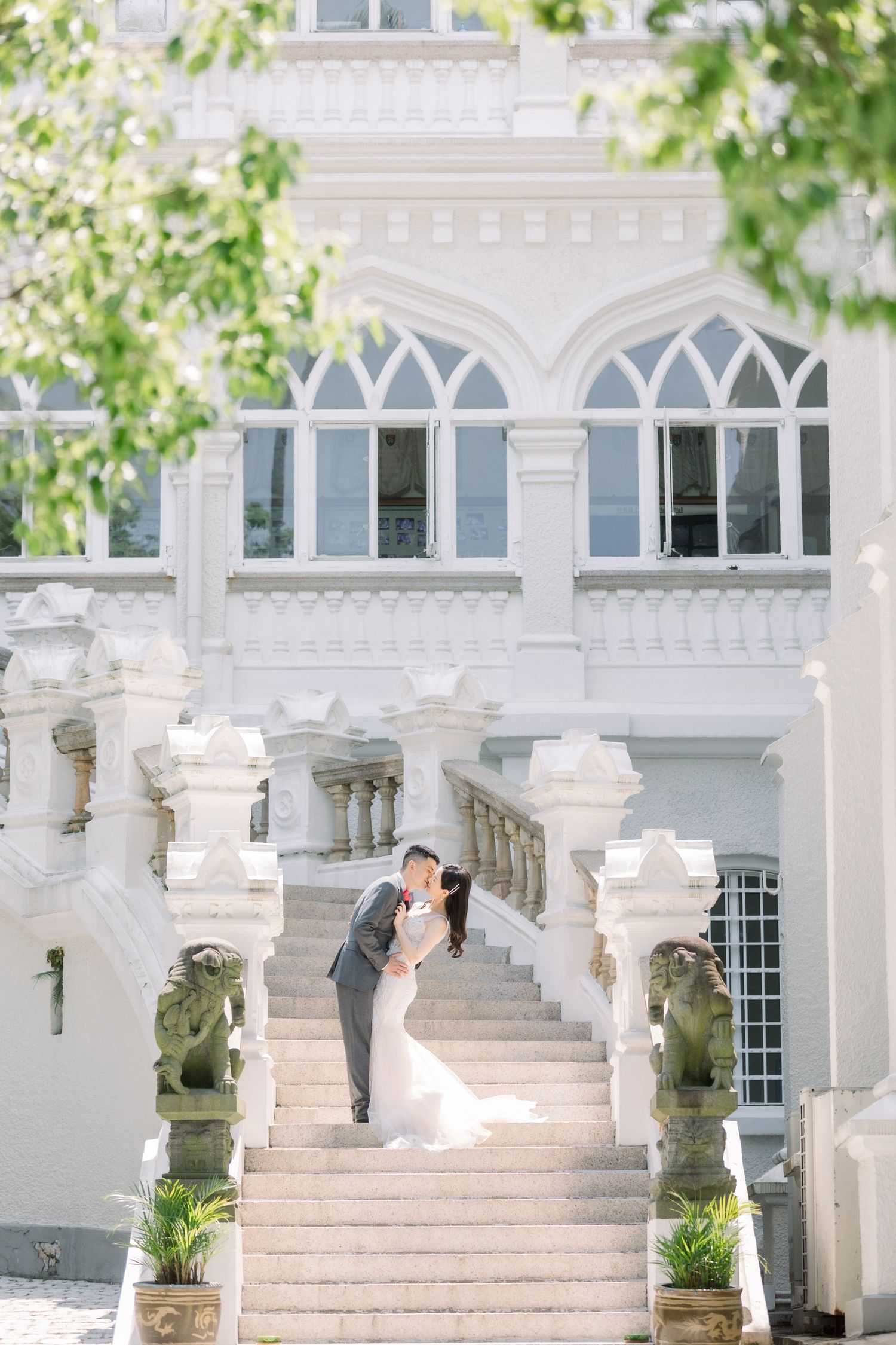 A couple stands on grand white marble staircase with ornate Gothic architecture and arched windows in the background.