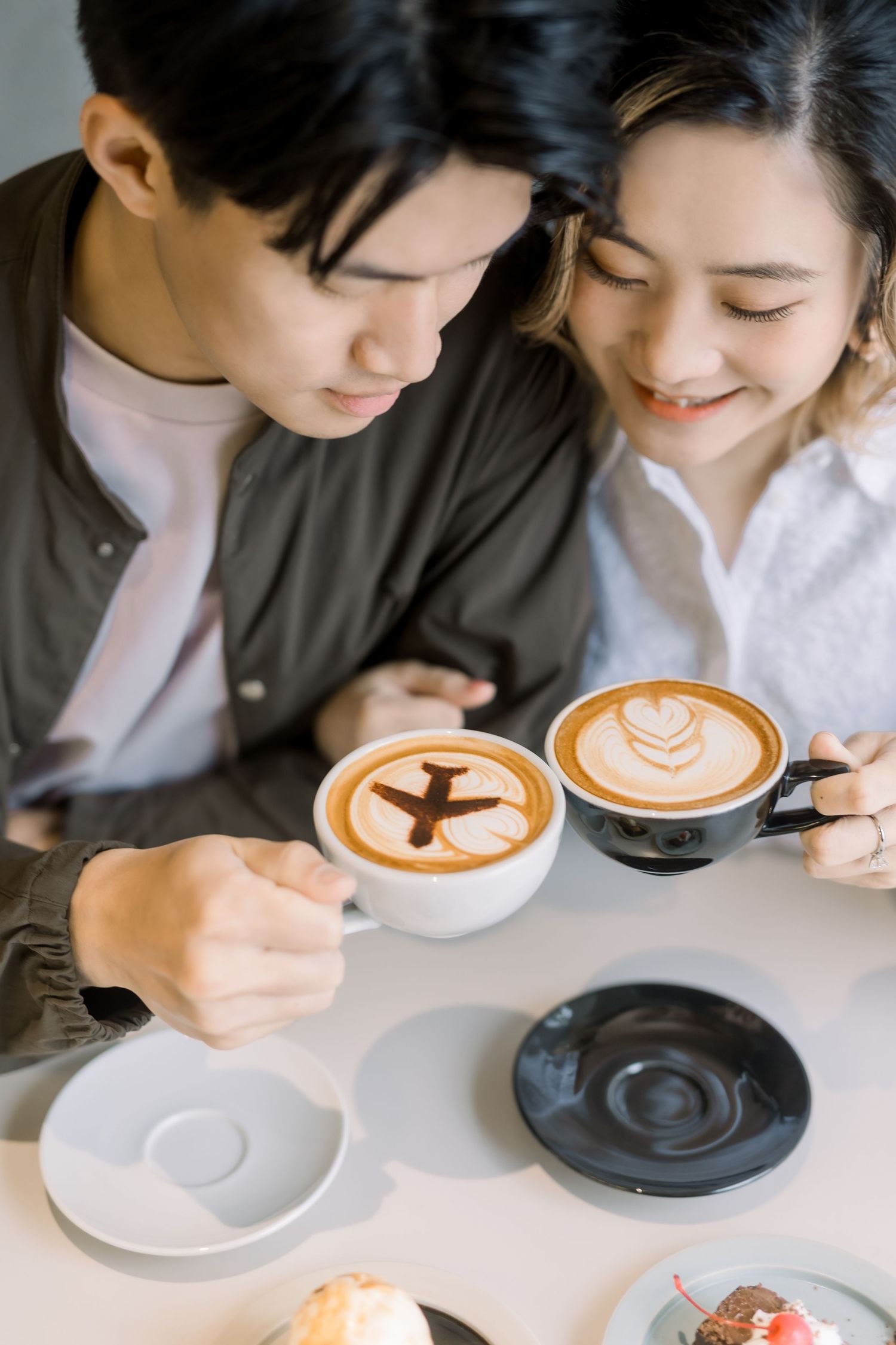Young Asian couple smiling and holding lattes with heart and airplane latte art at a cozy cafe.