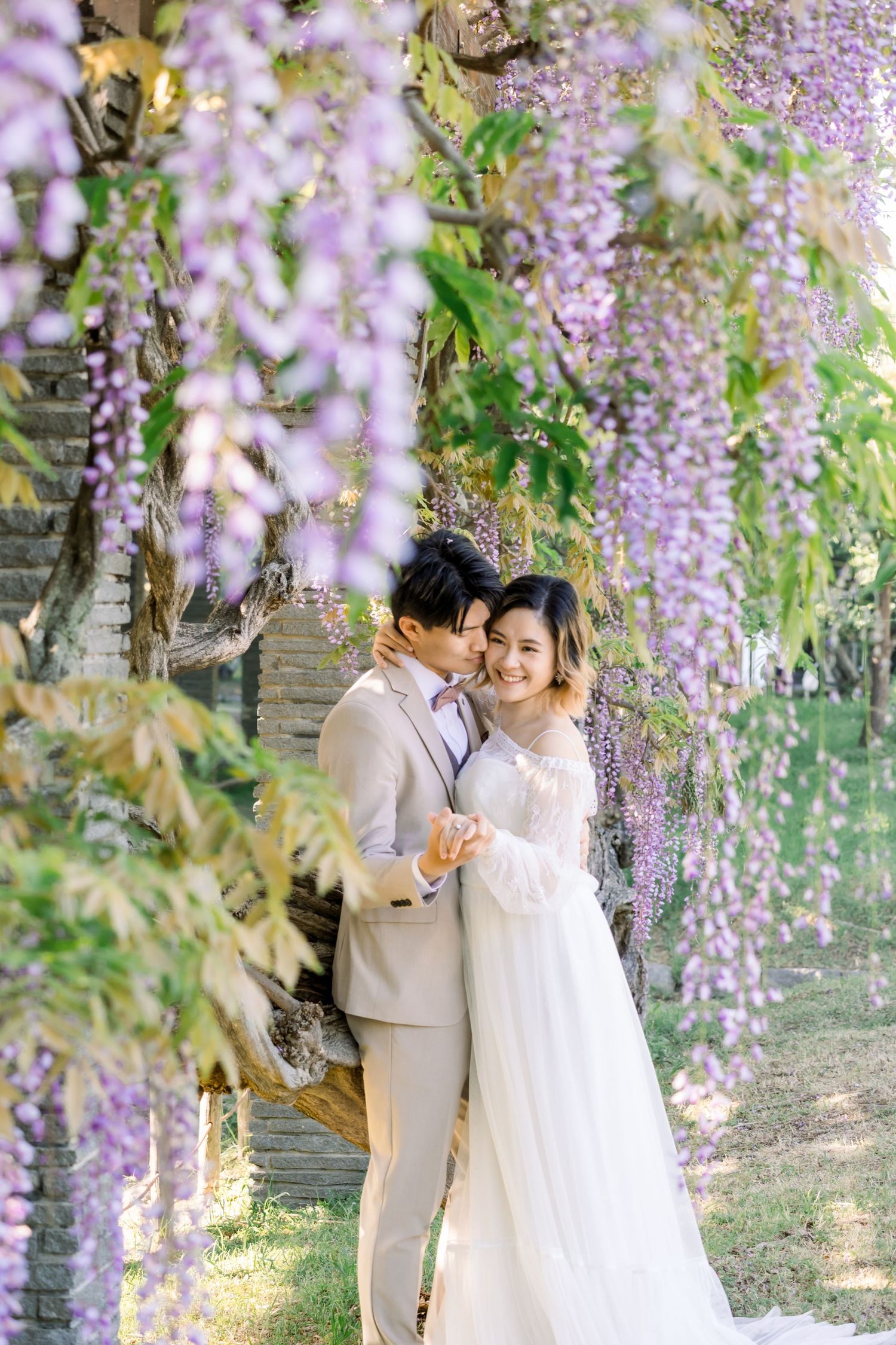 Couple in wedding attire embracing under cascading purple wisteria blooms in a lush garden setting.