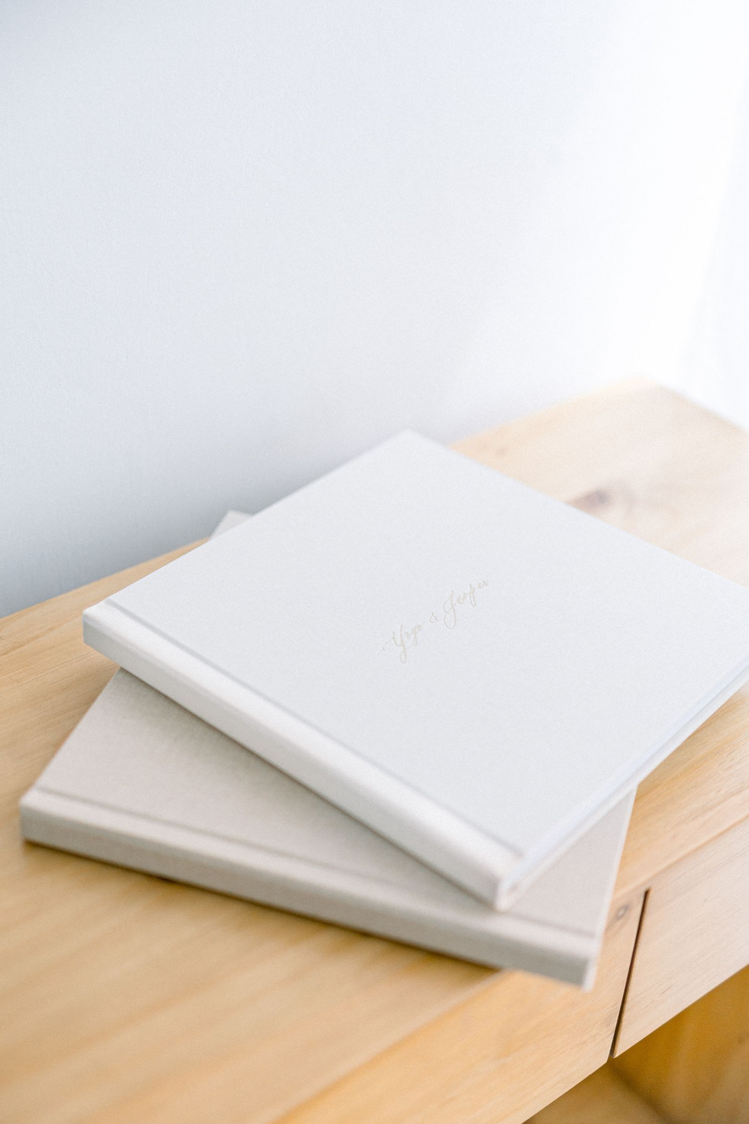 Two white hardcover photo albums stacked on a light wooden surface in a minimalist setting.