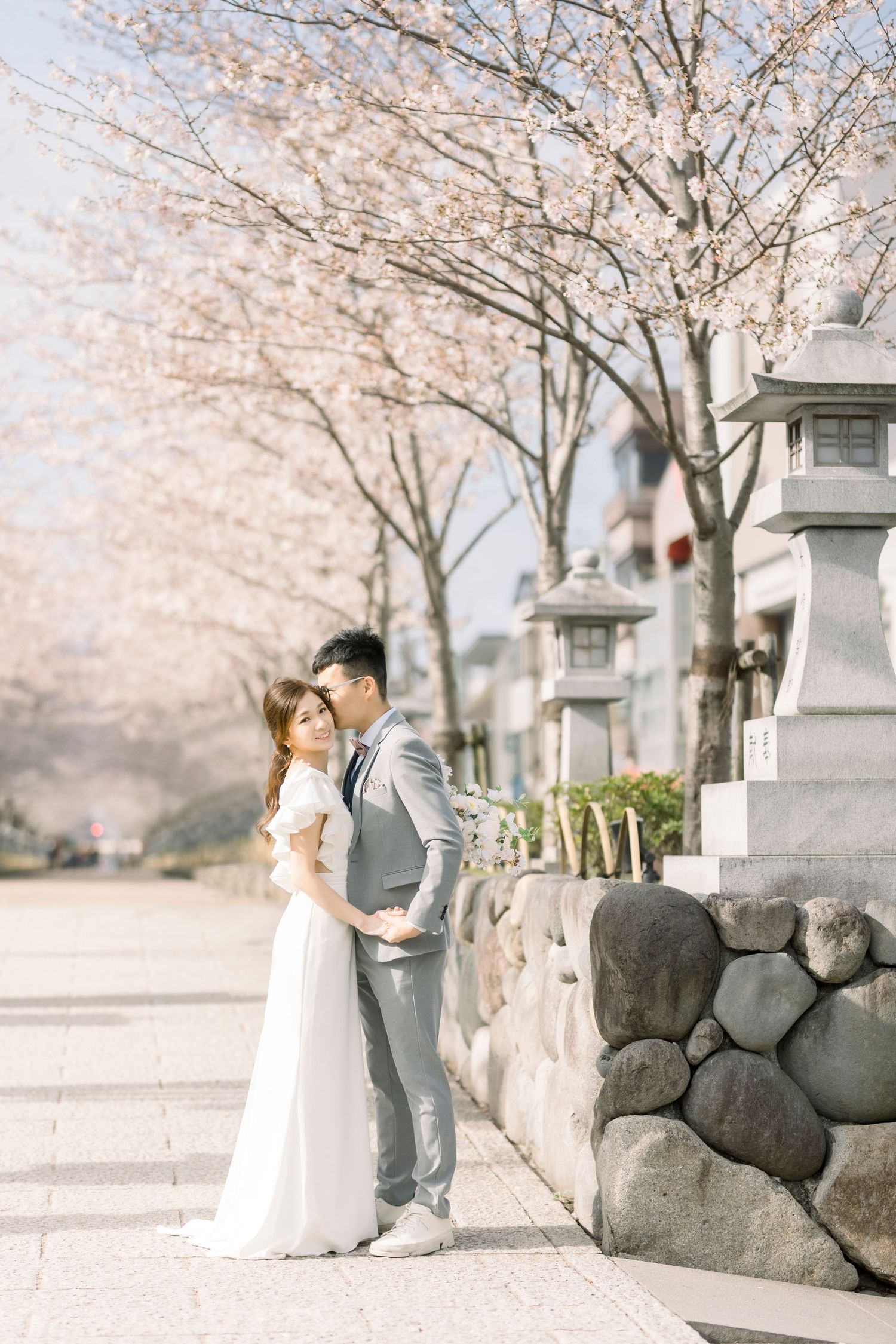 Couple in wedding attire embracing under blooming cherry blossom trees beside stone lanterns in Japan.