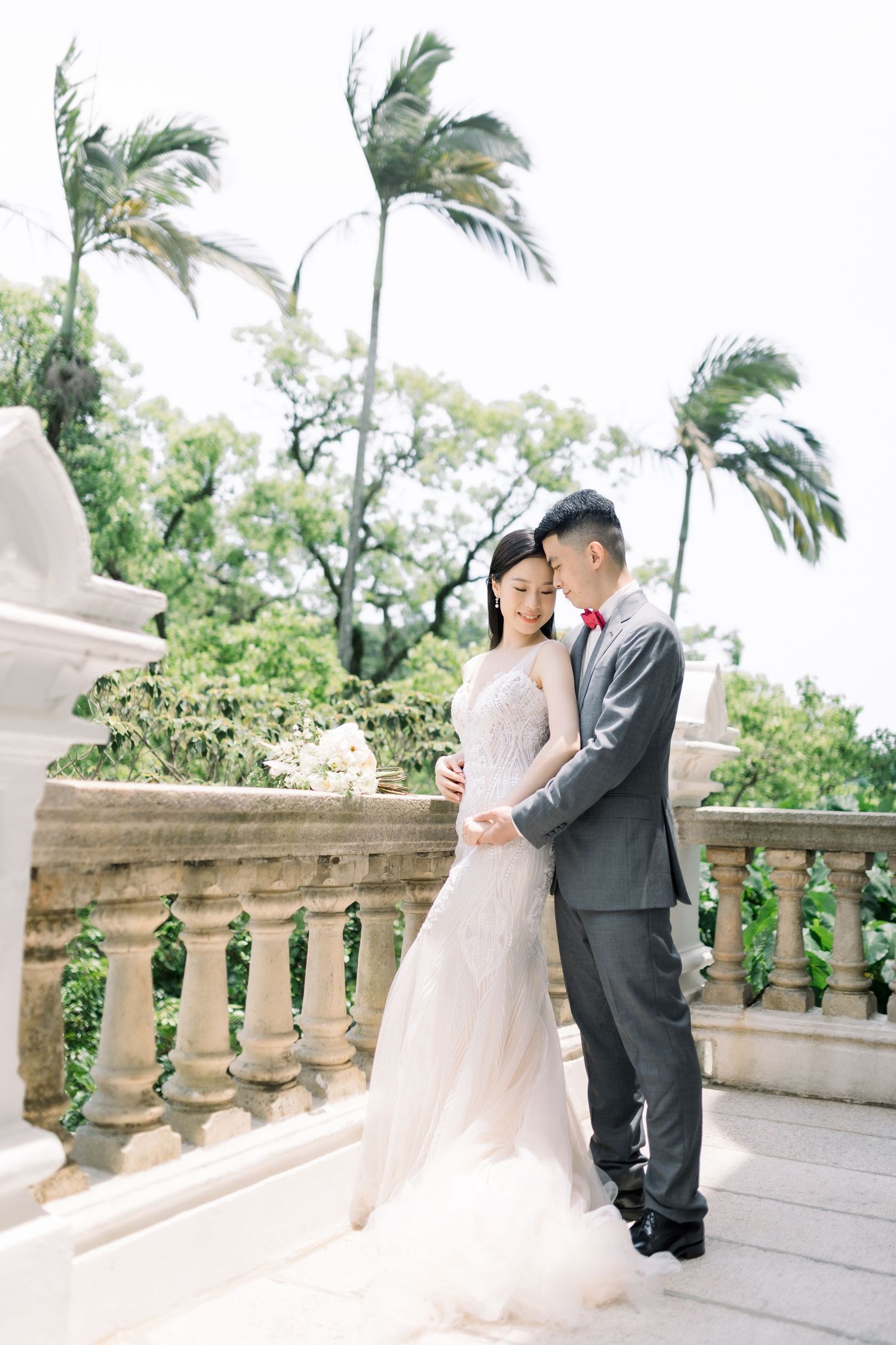 Couple in wedding attire embracing on stone balcony with palm trees, bride in white gown, groom in gray suit.