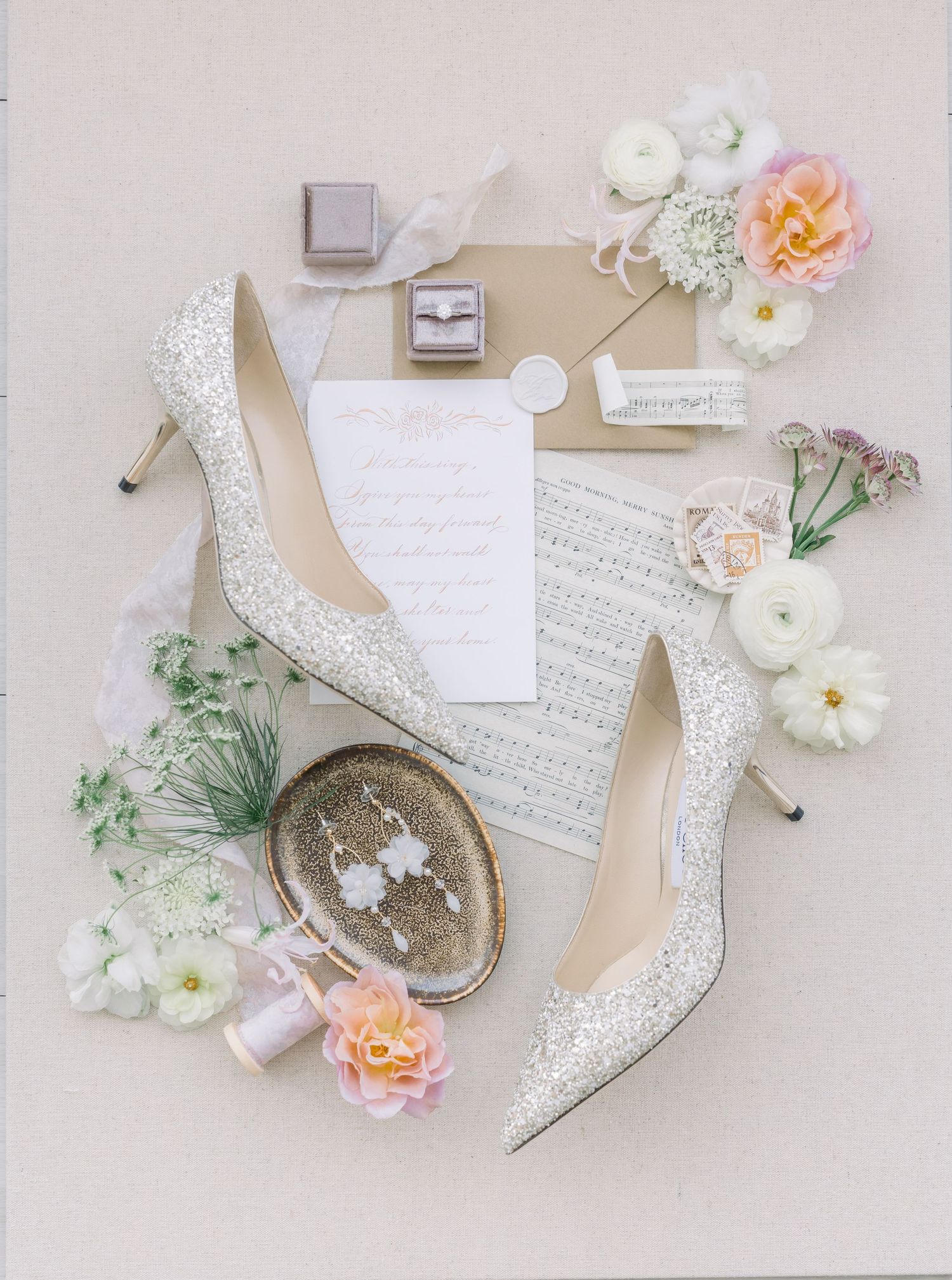 Elegant wedding details flatlay with white lace heels, invitations, flowers, and vintage accessories on light background.