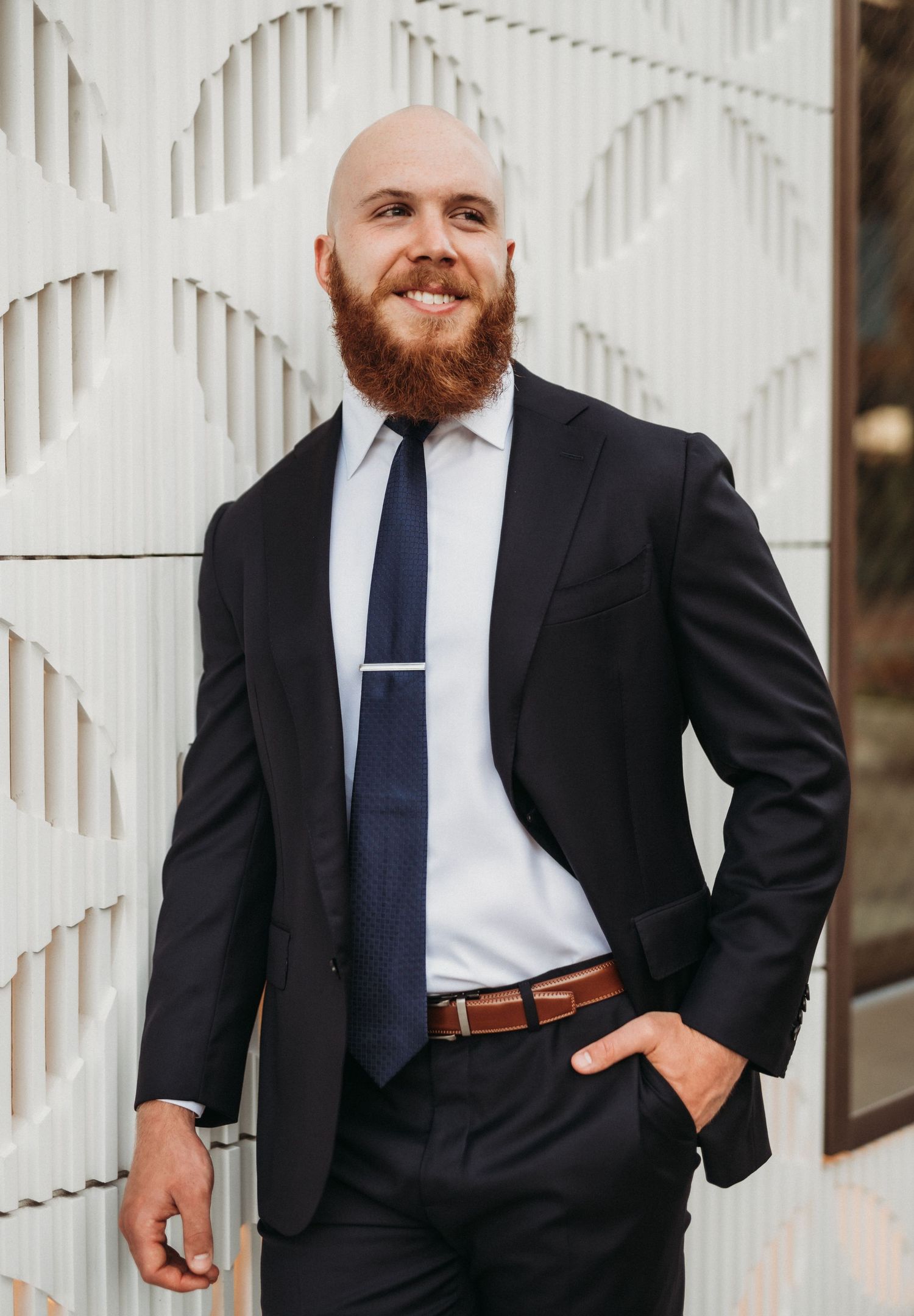 Professional portrait of someone in a classic black suit with navy tie and white shirt standing against textured wall pattern.