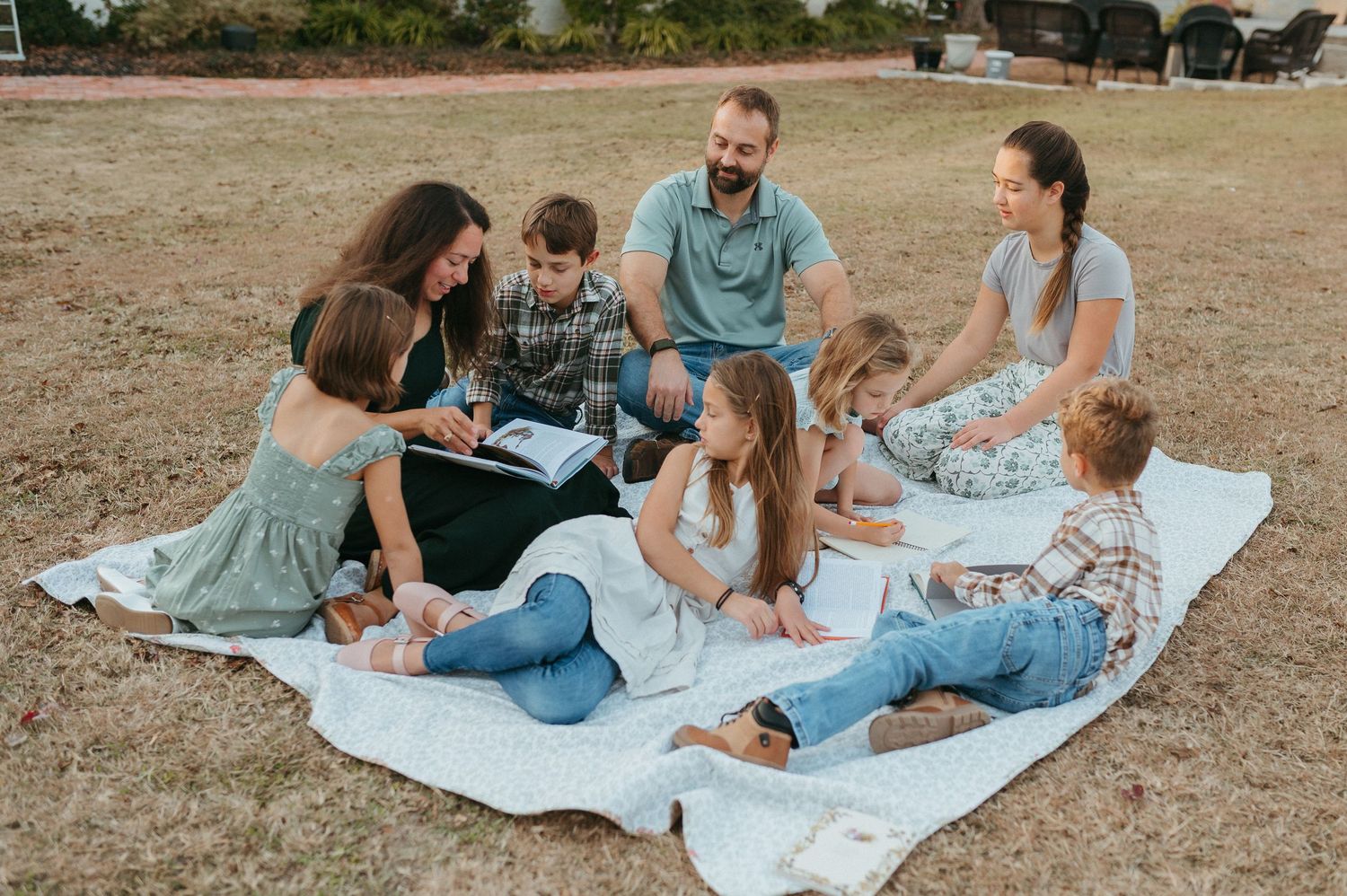 Family group sitting on white blanket outdoors on grass, reading and talking together on a sunny day.