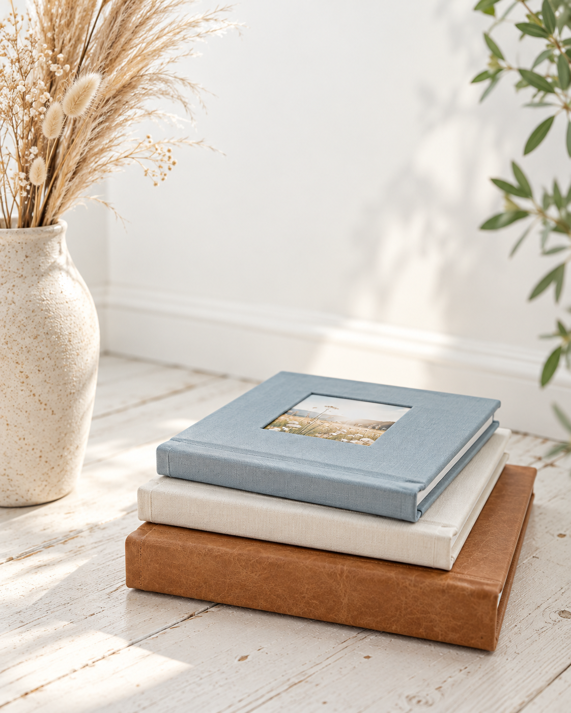 Stack of three photo albums in blue, cream and tan leather on a white wooden floor beside a textured vase of dried pampas grass.