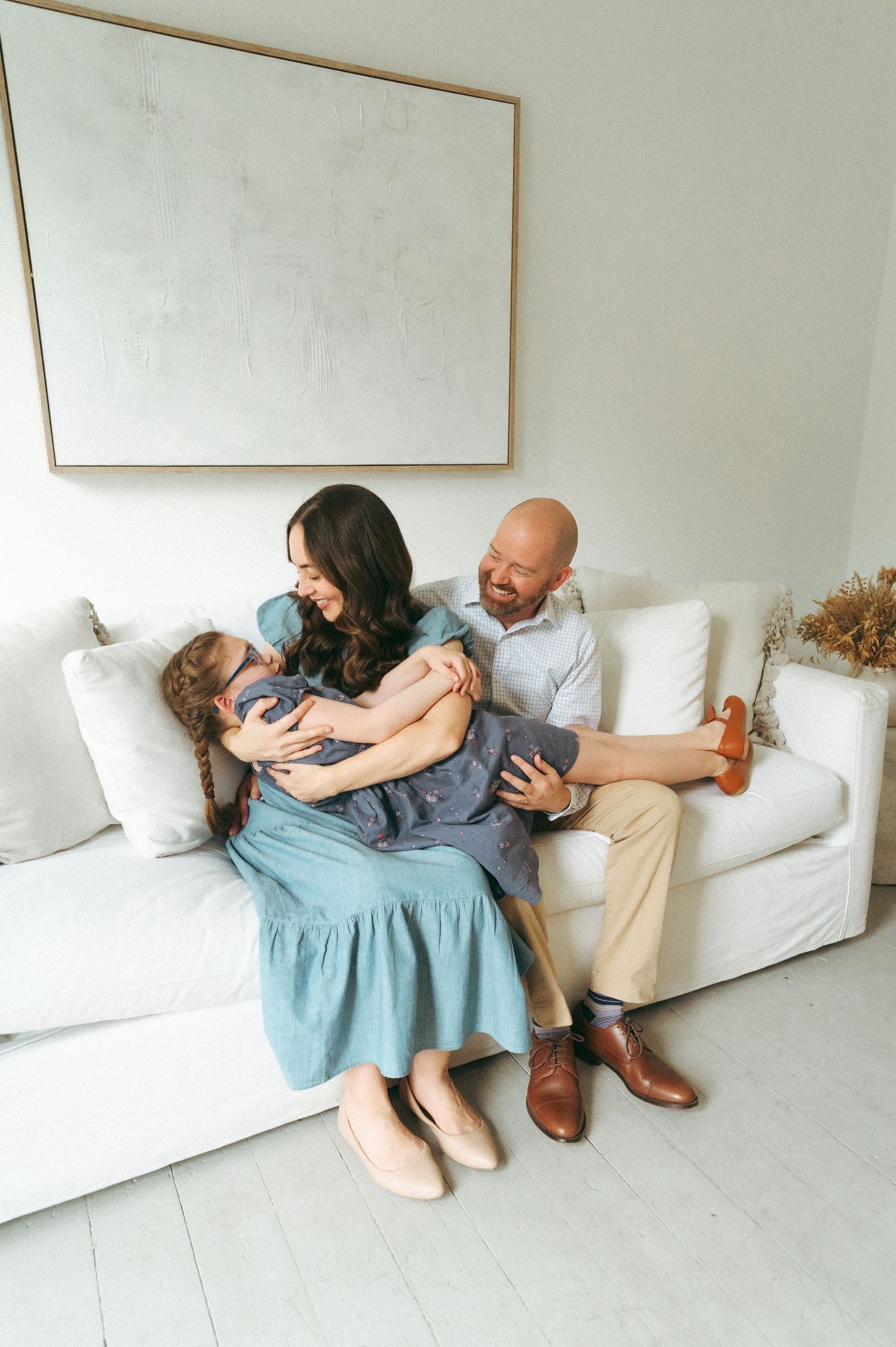 A happy family of three sitting on a white sofa, laughing and playing together in a bright, modern living room.