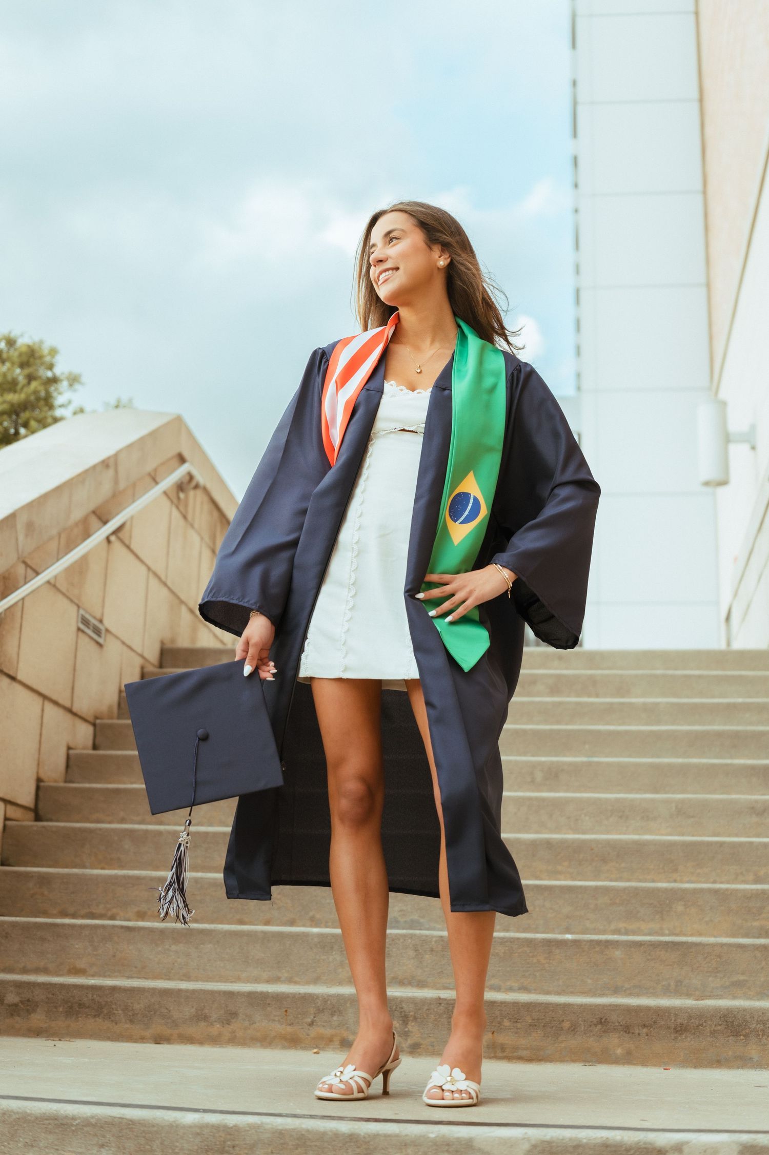 Young woman in graduation gown holding cap, wearing Brazilian flag stole, smiling on outdoor steps.