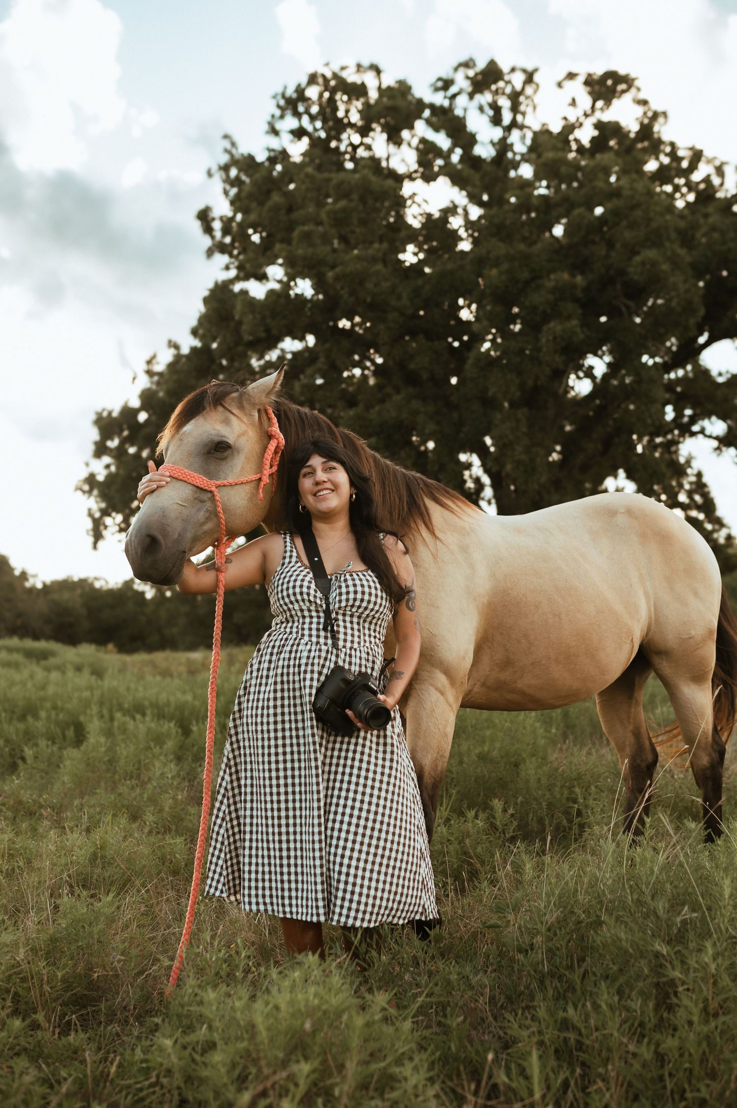 A woman in a gingham dress stands beside a cream-colored horse in a grassy field on an overcast day.