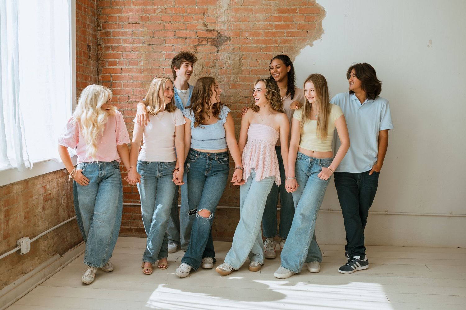 Group of friends in coordinated denim and blue outfits lean against brick wall while laughing together.