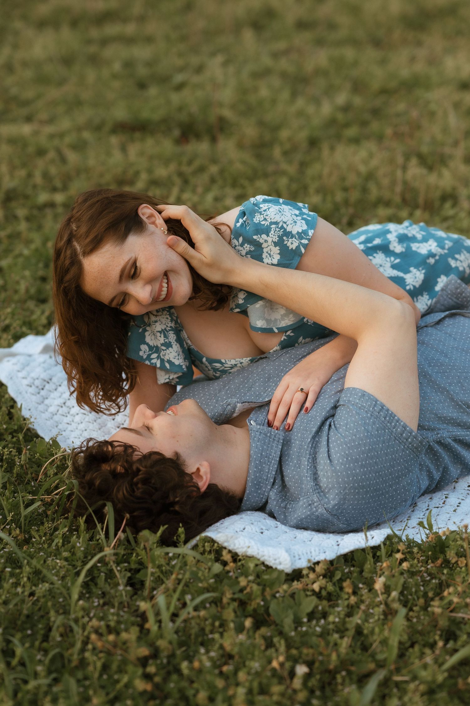 A smiling couple lying on a blanket in green grass, sharing a romantic moment outdoors.