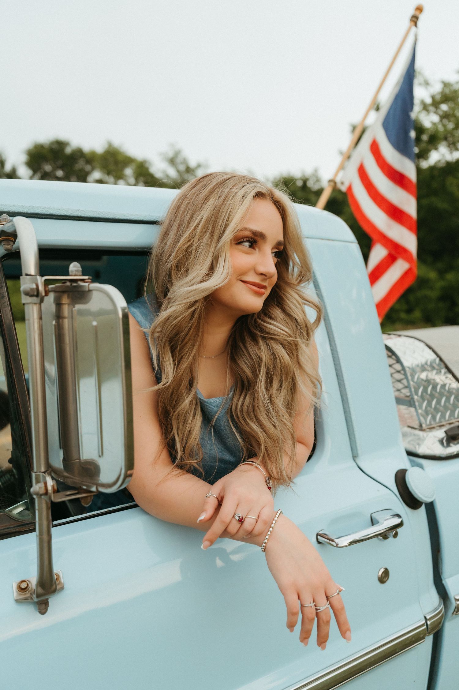 Blonde woman leaning out of a vintage light blue truck window with an American flag in the background.