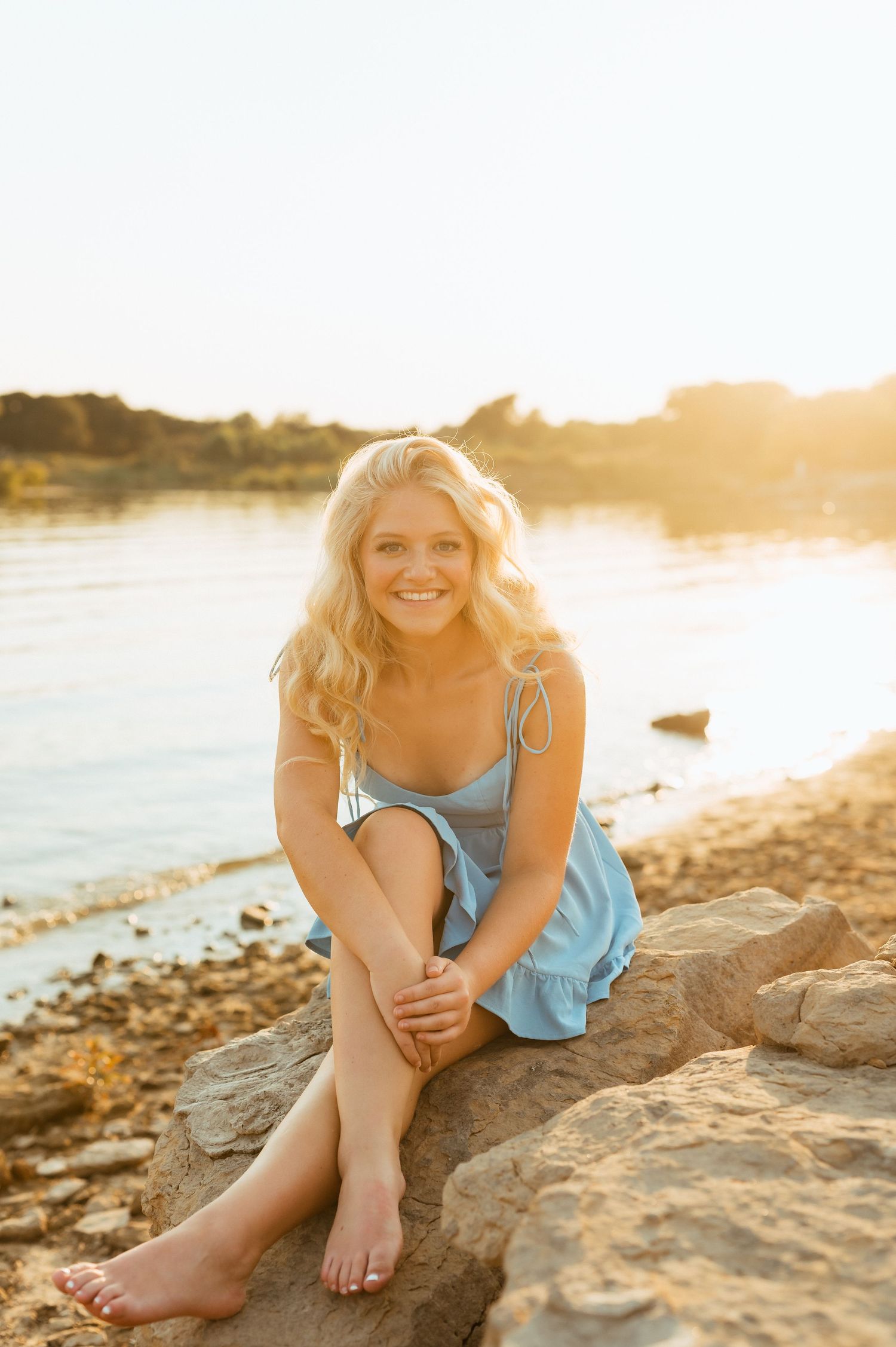 Smiling blonde woman in light blue sundress sitting on rocks by a sunlit lake shore.