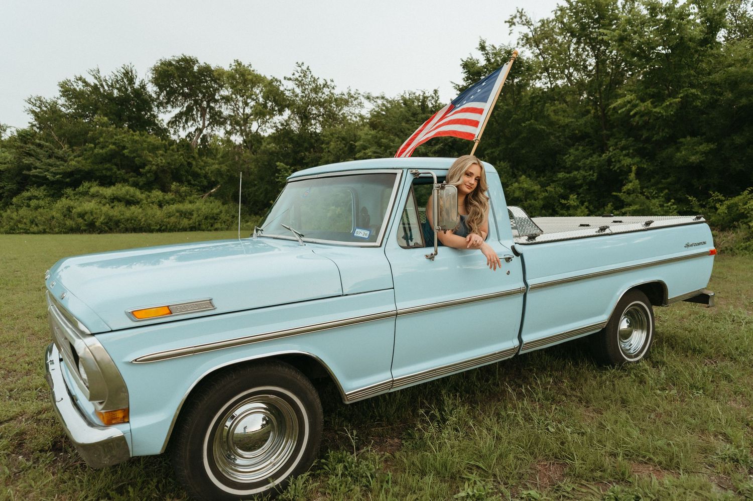 Woman leaning out of a classic light blue Ford pickup truck holding an American flag in a green field.