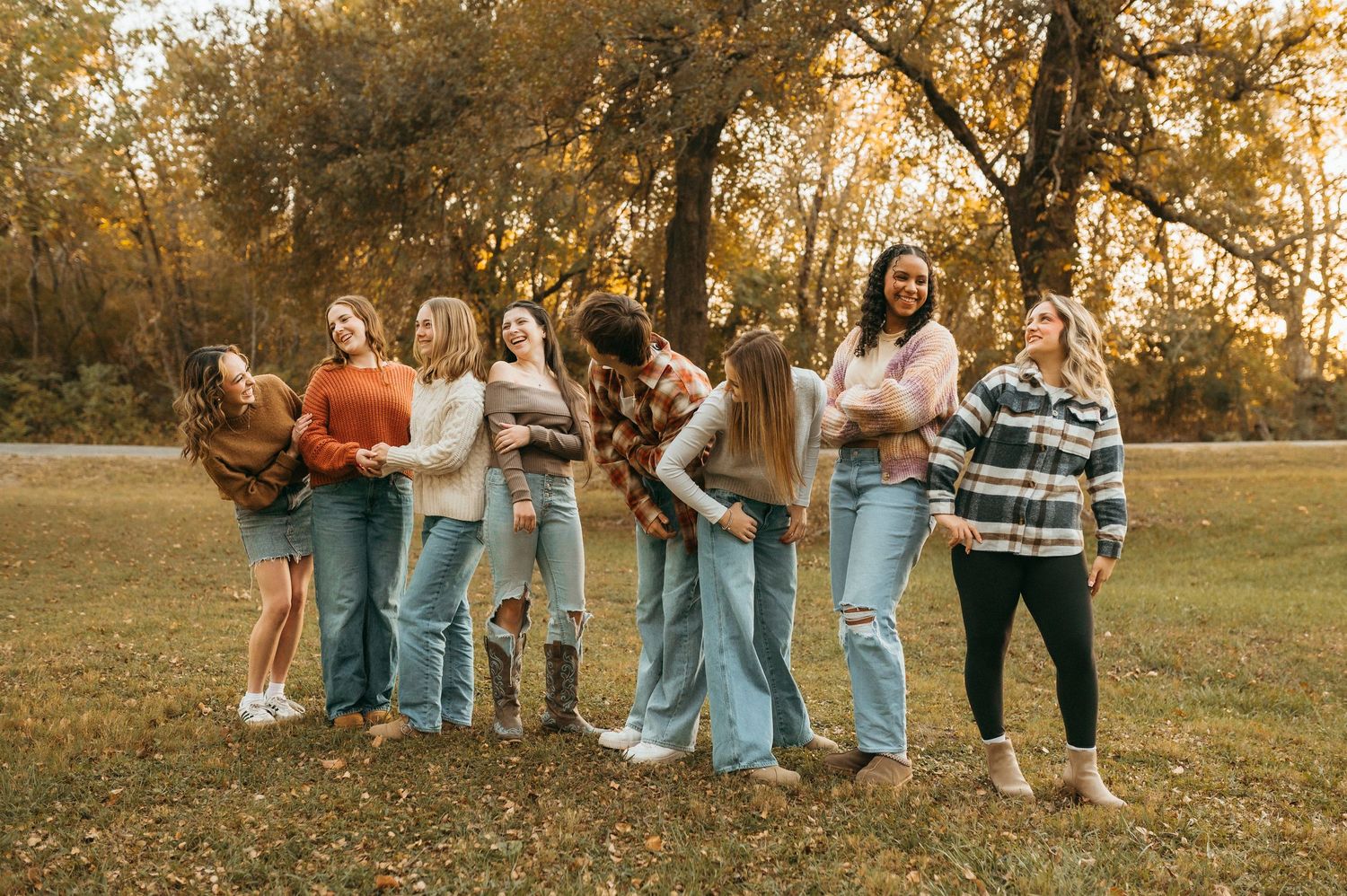 Group of friends laughing together in a park during golden hour autumn afternoon wearing casual denim and sweaters.
