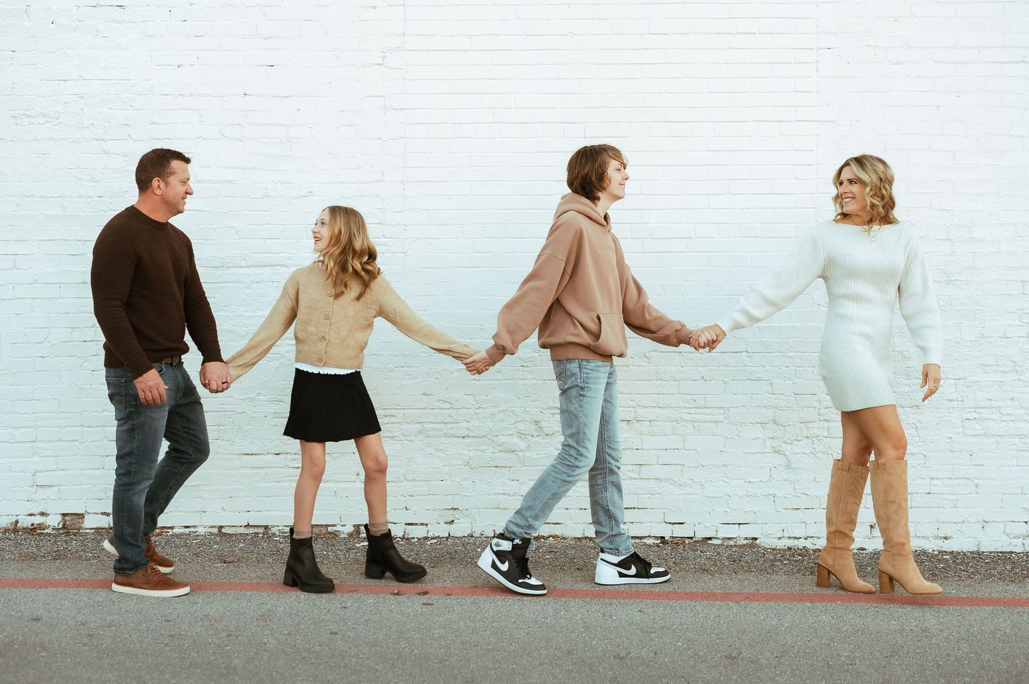 Family of four holding hands in a line against a white brick wall outdoors.