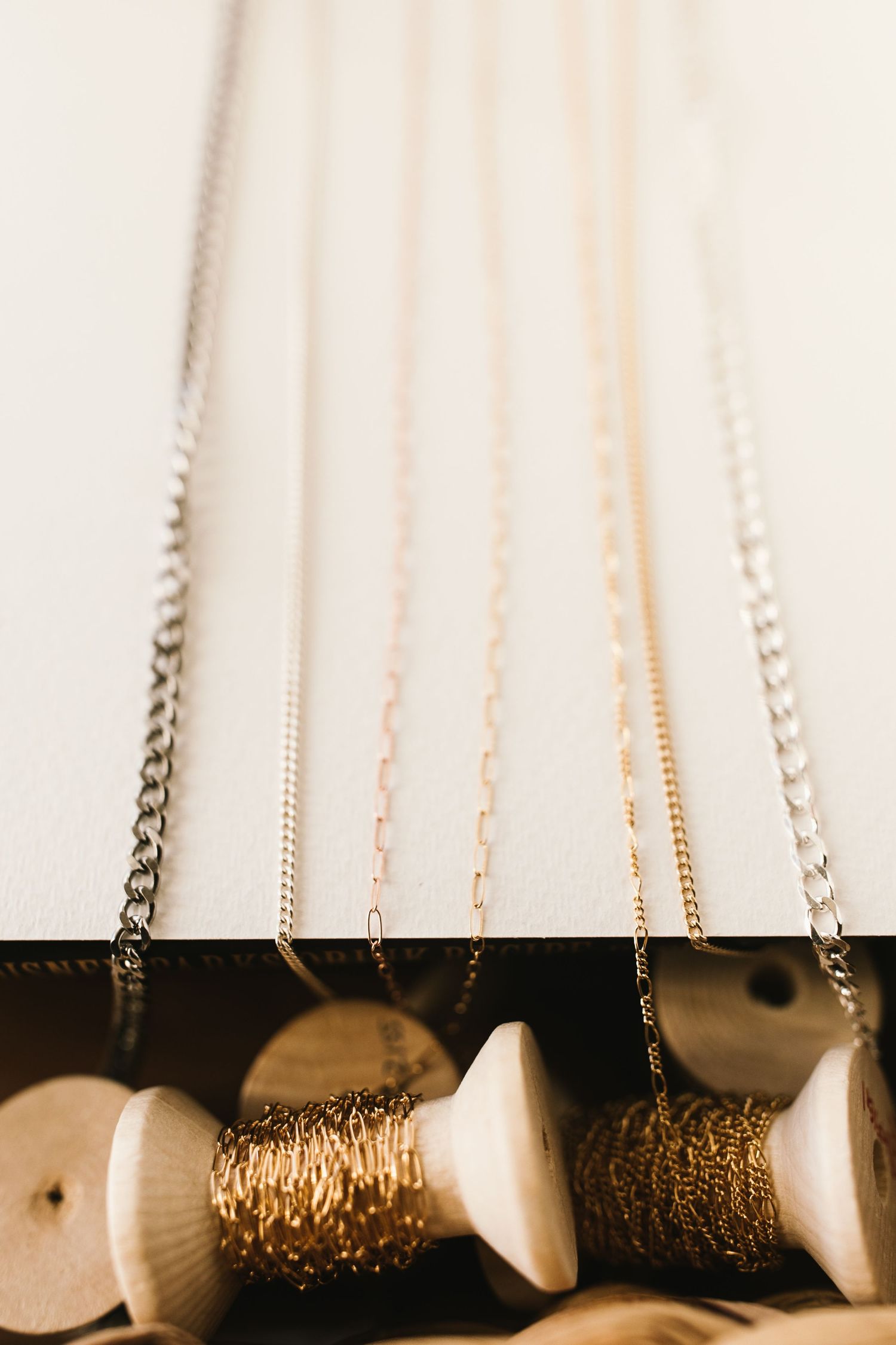 Multiple metal chains in gold and silver hanging from wooden spools against a white background.