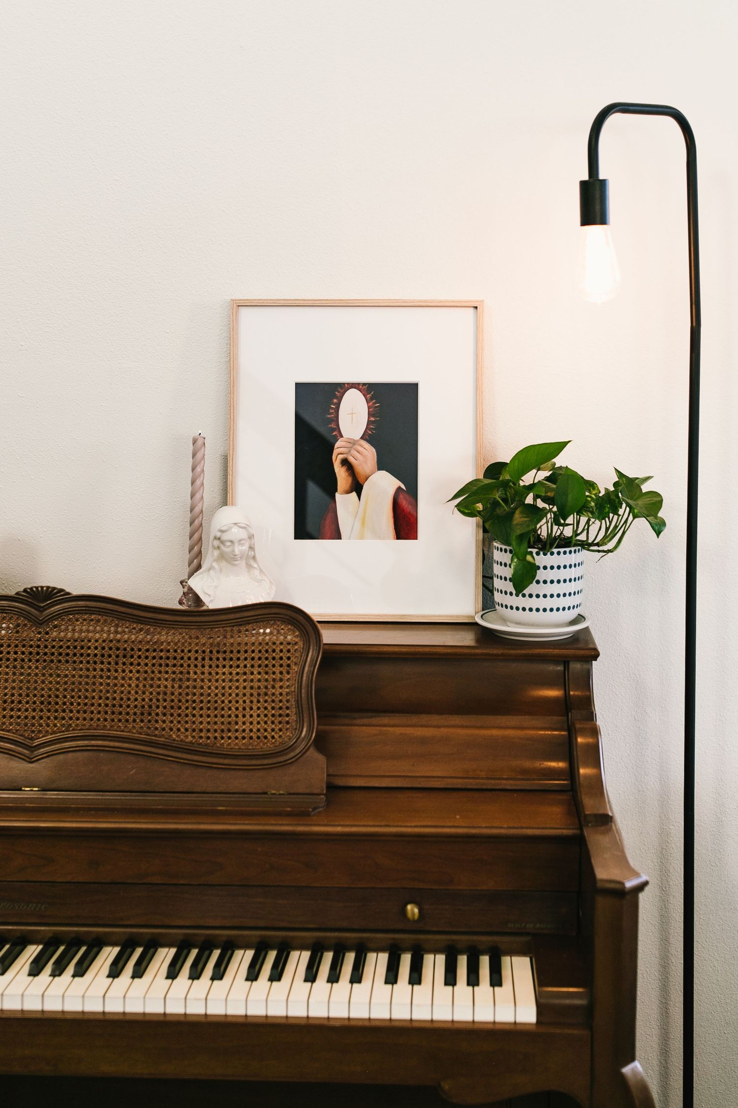 A cozy piano corner with a framed artwork, modern floor lamp, and potted plant atop a vintage upright piano.