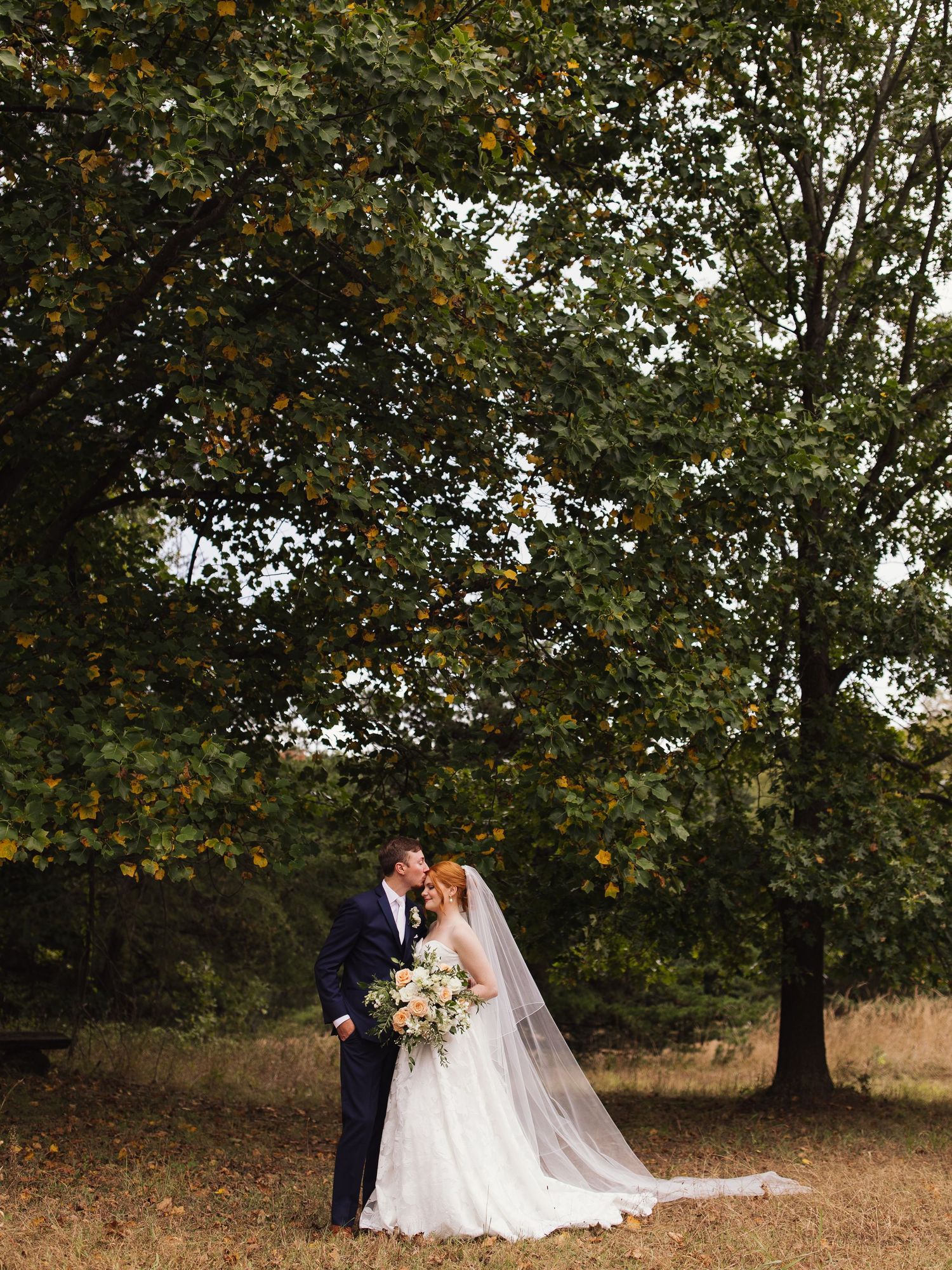 A couple poses for romantic wedding photos under autumn trees in a scenic outdoor setting.