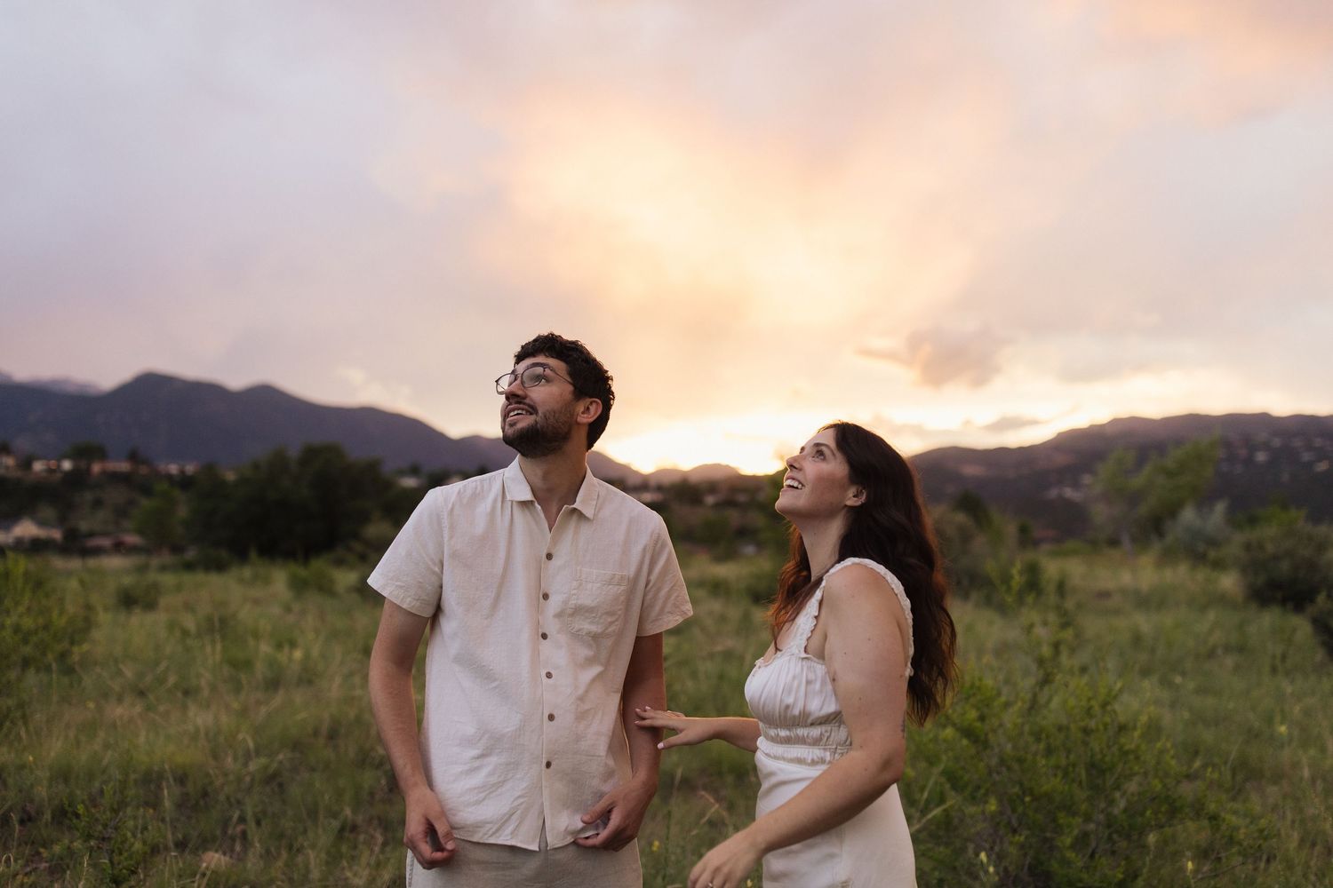 Couple enjoying a romantic sunset moment in a scenic grassland field with mountains in the background.