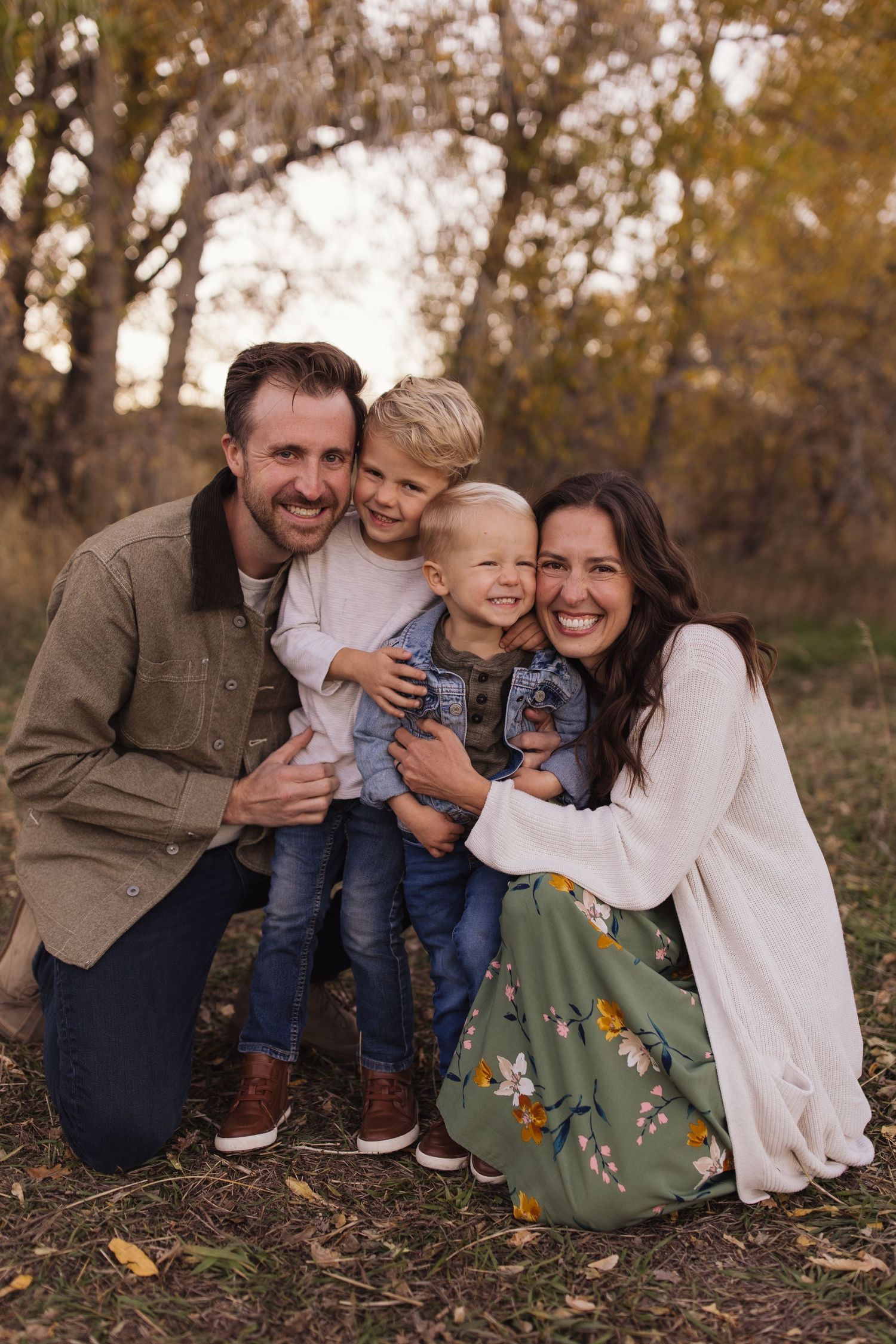 A family poses together outdoors in autumn with fallen leaves and trees in the background.