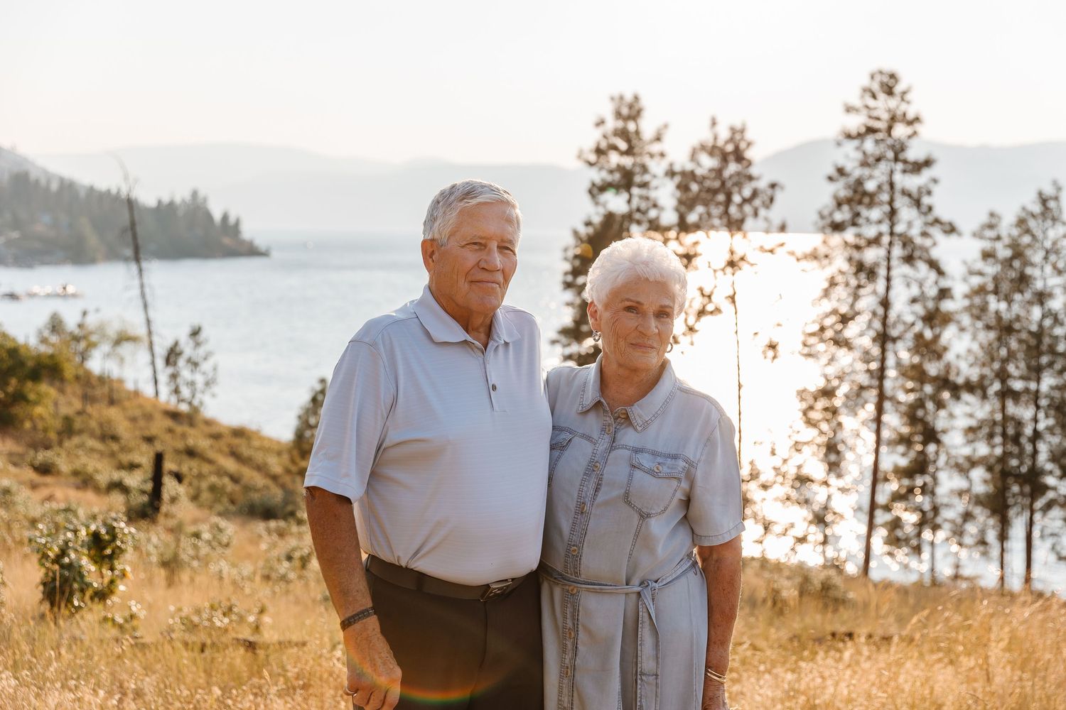 Elderly couple stands together on grassy hillside overlooking scenic lake and evergreen trees at sunset.