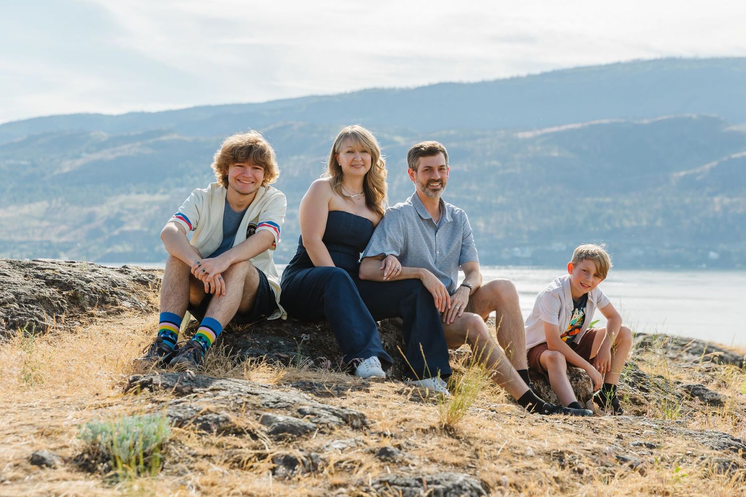 A family sits together on a rocky outcrop overlooking a scenic lake with mountains in the background on a sunny day.