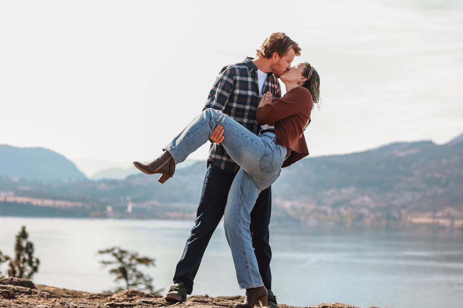 A couple shares a romantic dip kiss on a scenic mountain overlook with a lake view in the background.