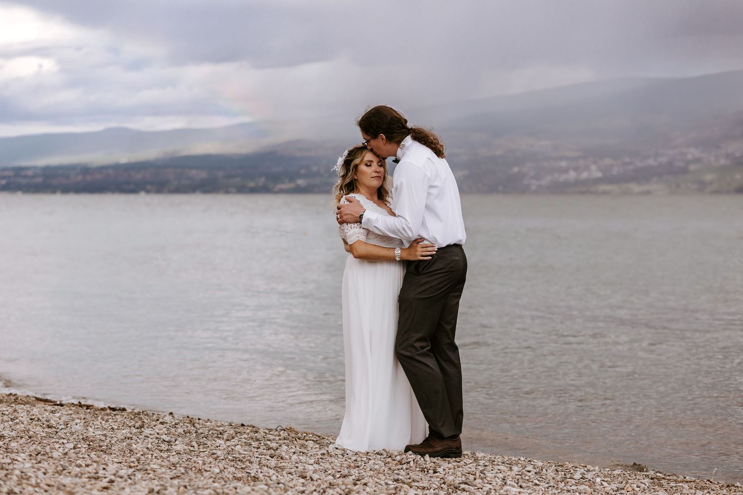 A couple shares a romantic embrace on a rocky beach with mountains and cloudy skies in the background.