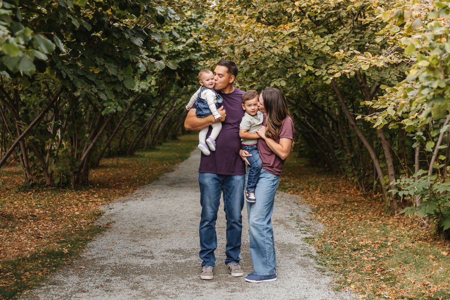 A couple walks down a tree-lined autumn path while holding their dogs on a peaceful afternoon.