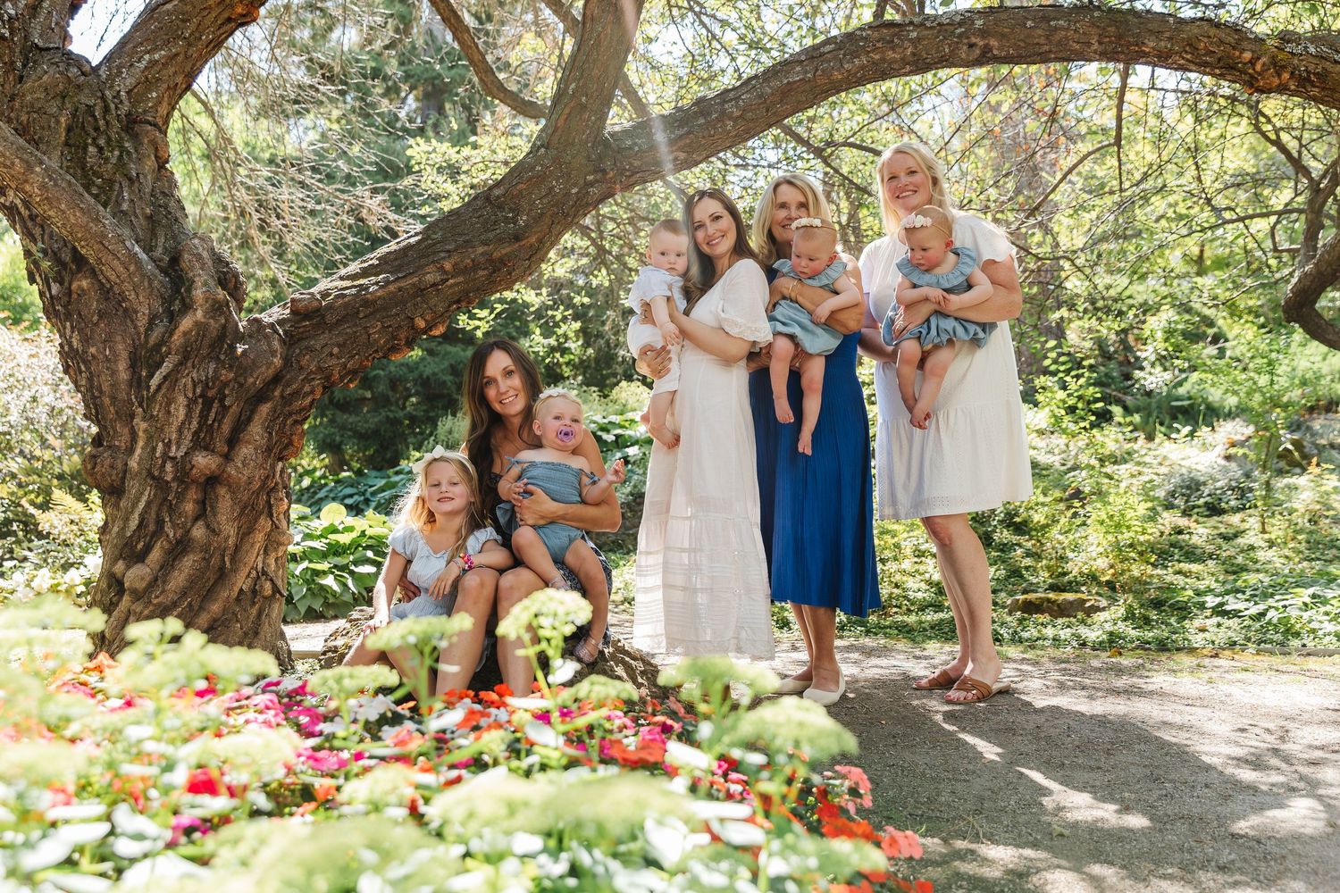 Extended family gathering under a majestic tree branch in a beautiful garden setting with red flowers.