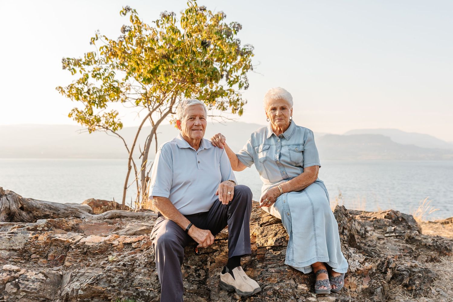 Senior couple sits together on rocky outcrop with ocean view and small tree during golden hour.