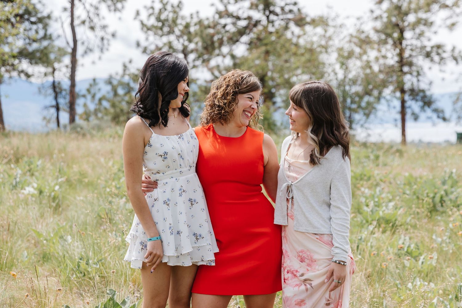 Three women in coordinated white and red outfits laugh together while walking through a sunny summer meadow.